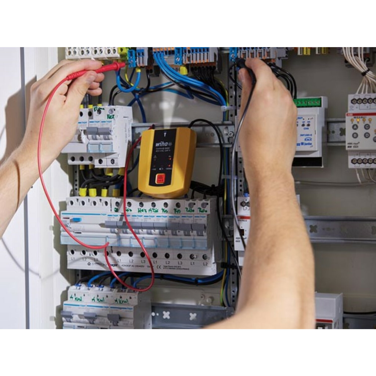 Elektriker testet Drähte in einem Schaltschrank mit roten und schwarzen Sonden, gelbes Gerät mittig.
