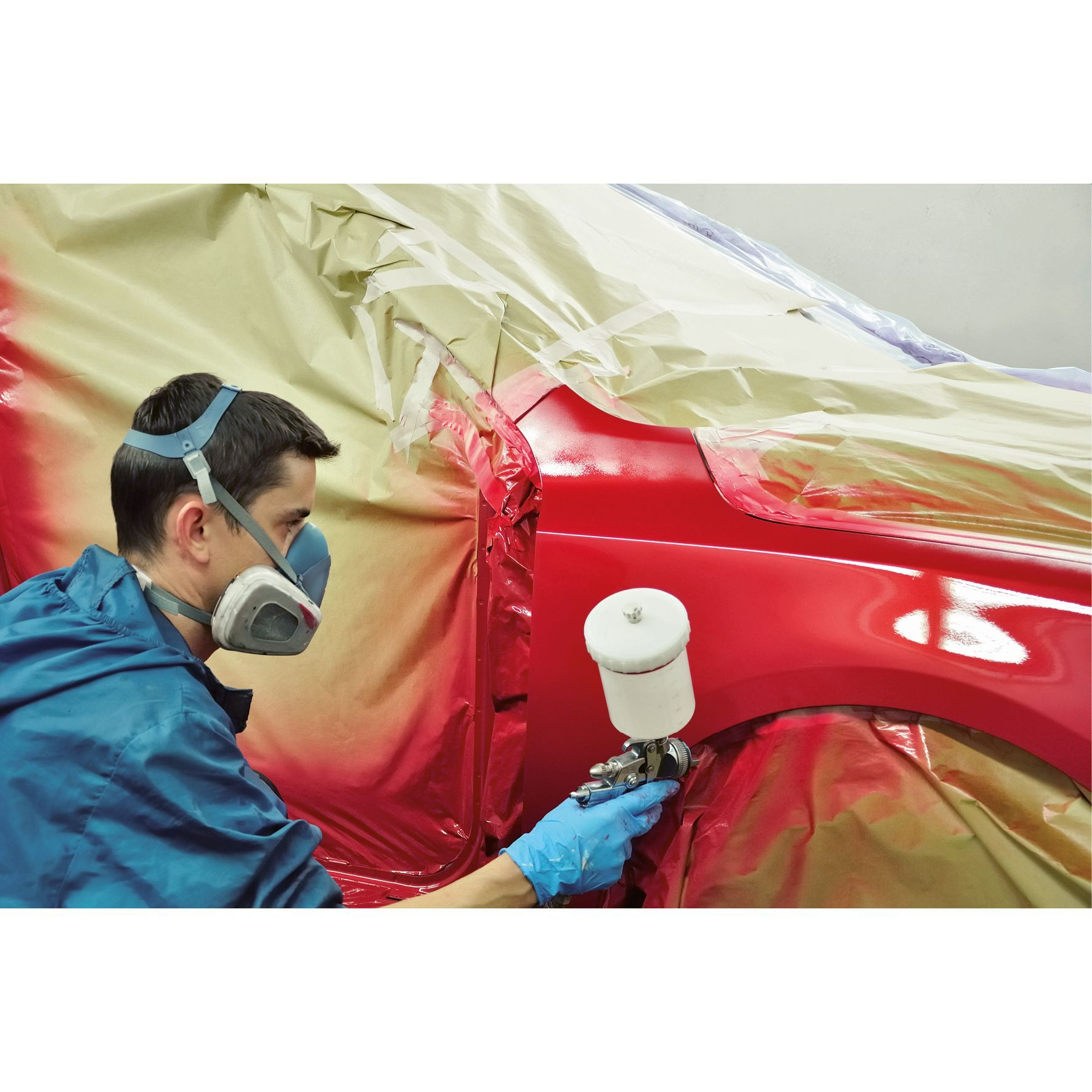 Persona con tuta blu che spruzza vernice su auto rossa, con respiratore.