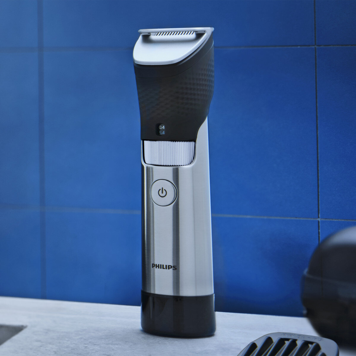 Srebrno-czarna maszynka do strzyżenia brody Philips na niebieskiej ścianie z kafelkami. Maszynka stoi na szarej powierzchni.