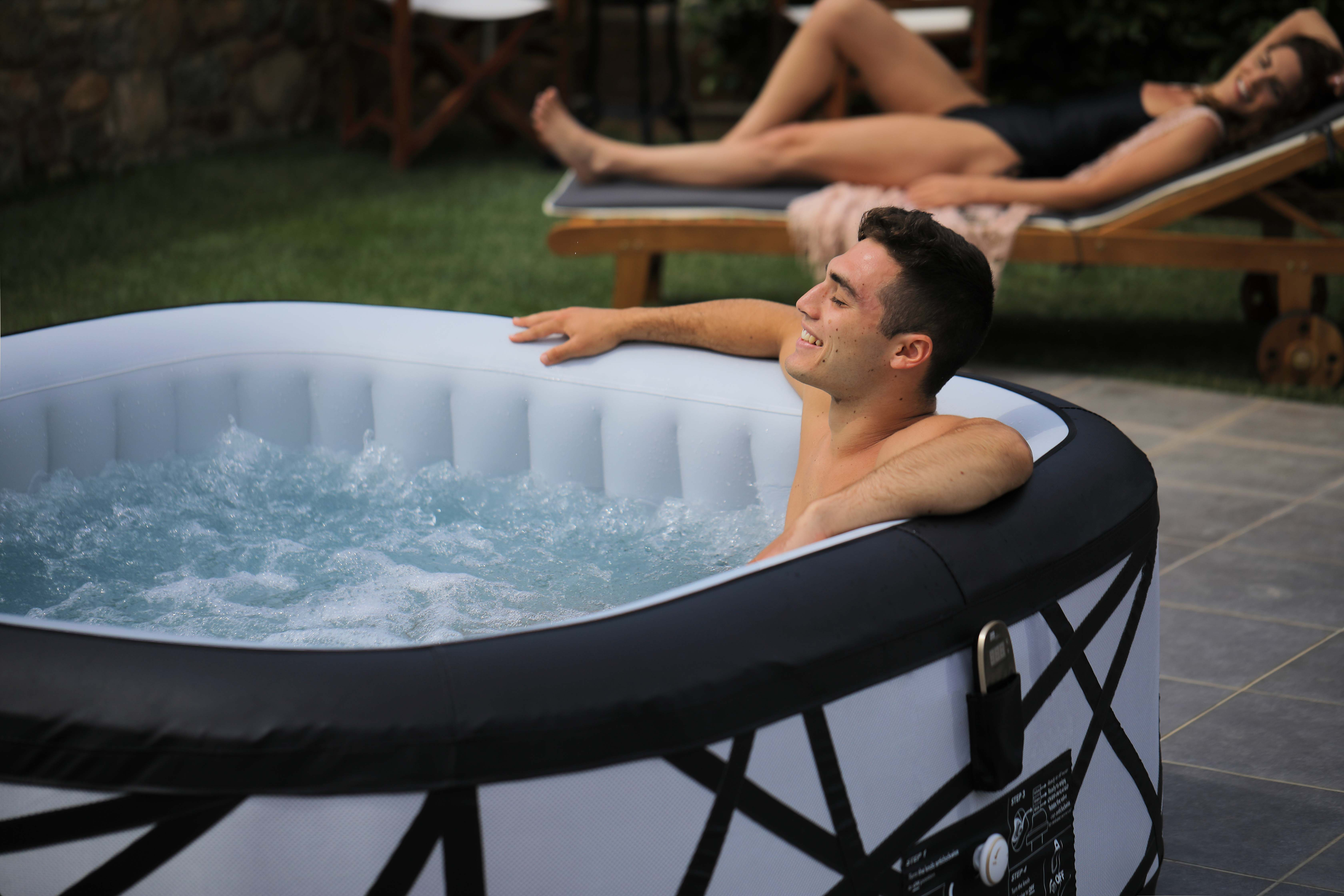 Ein Mann in einem Whirlpool lächelt. Eine Frau entspannt sich im Hintergrund auf einer Liege.