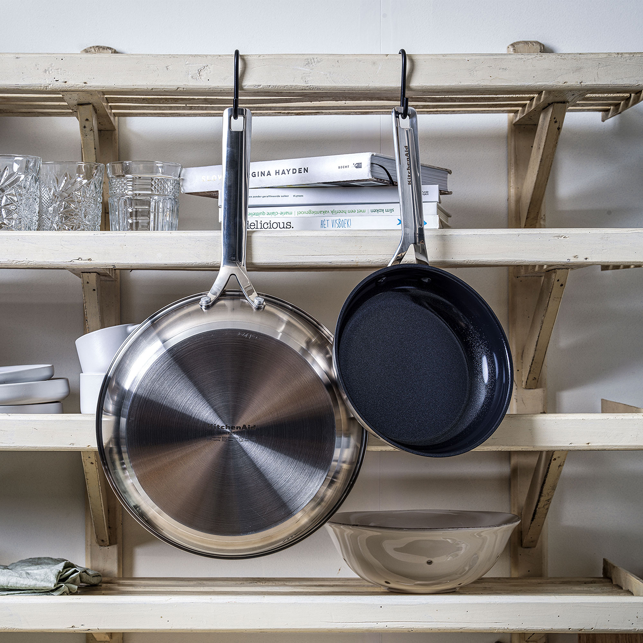Edelstahlpfanne und schwarze Pfanne hängen an einem Holzregal mit Büchern, Gläsern und Schalen sichtbar.