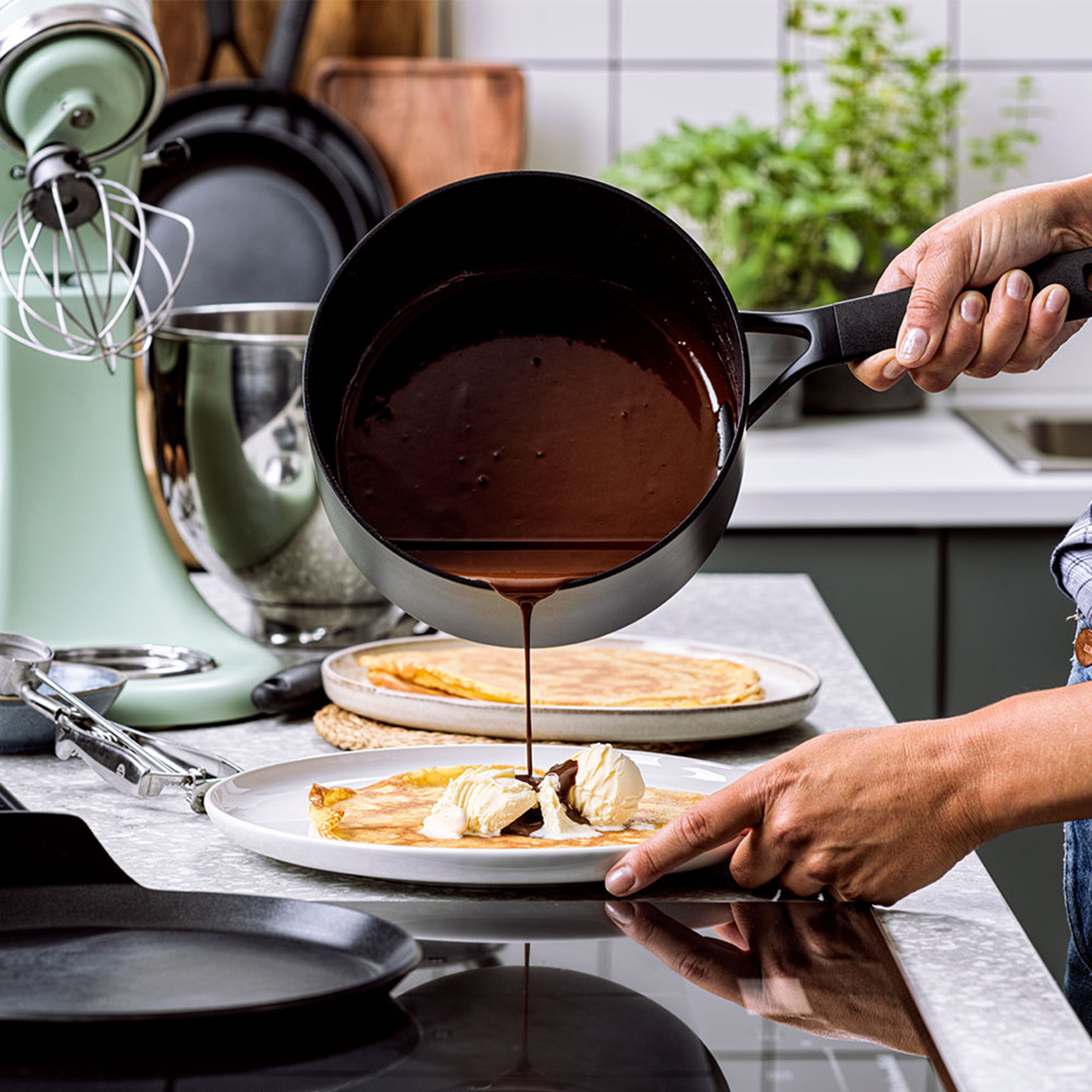 Hände gießen Schokoladensauce über Crêpe mit Eis. Küchenumgebung mit Geräten im Hintergrund.