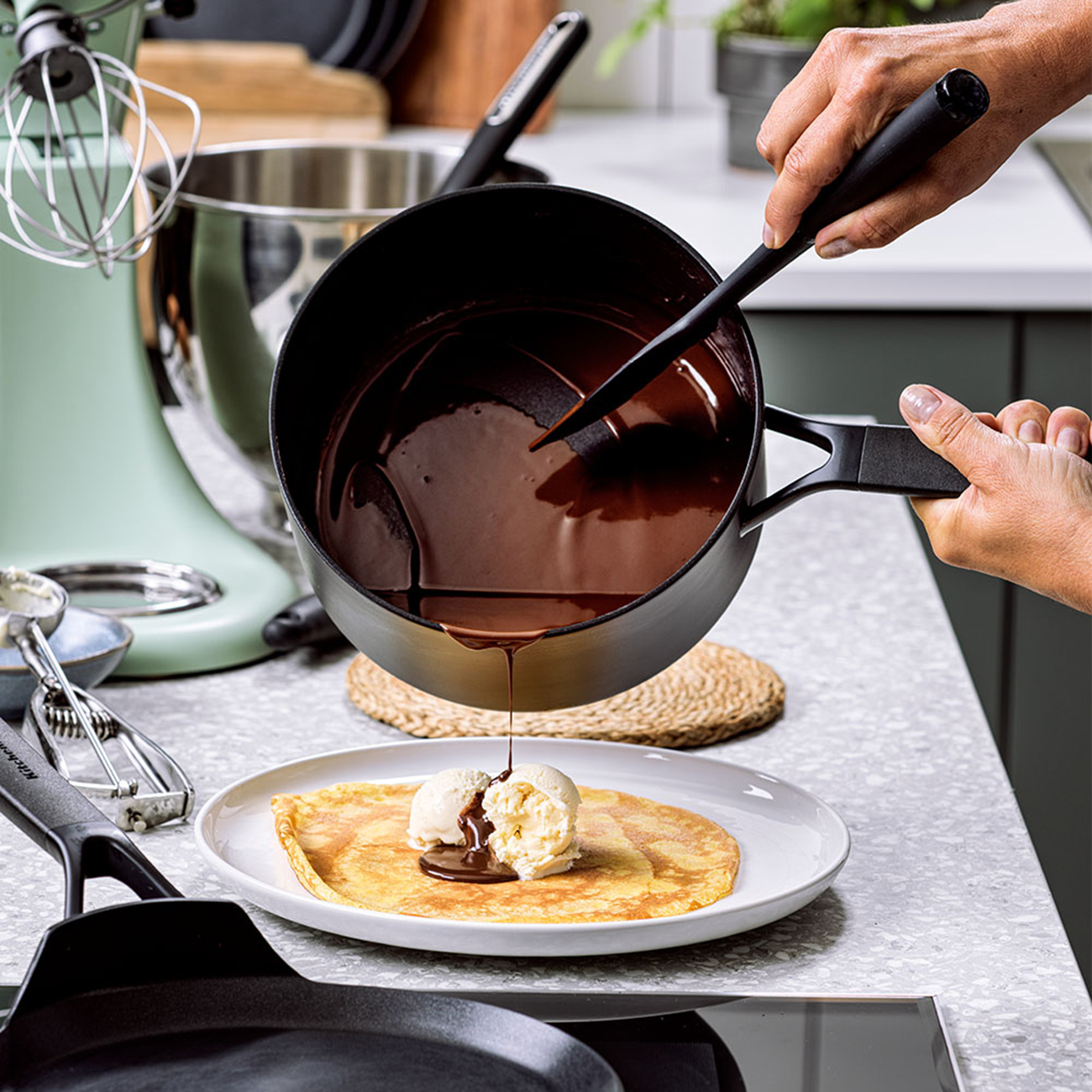 Schokoladensauce wird auf einen Crêpe mit Eis gegossen. Küchenutensilien sind im Hintergrund.