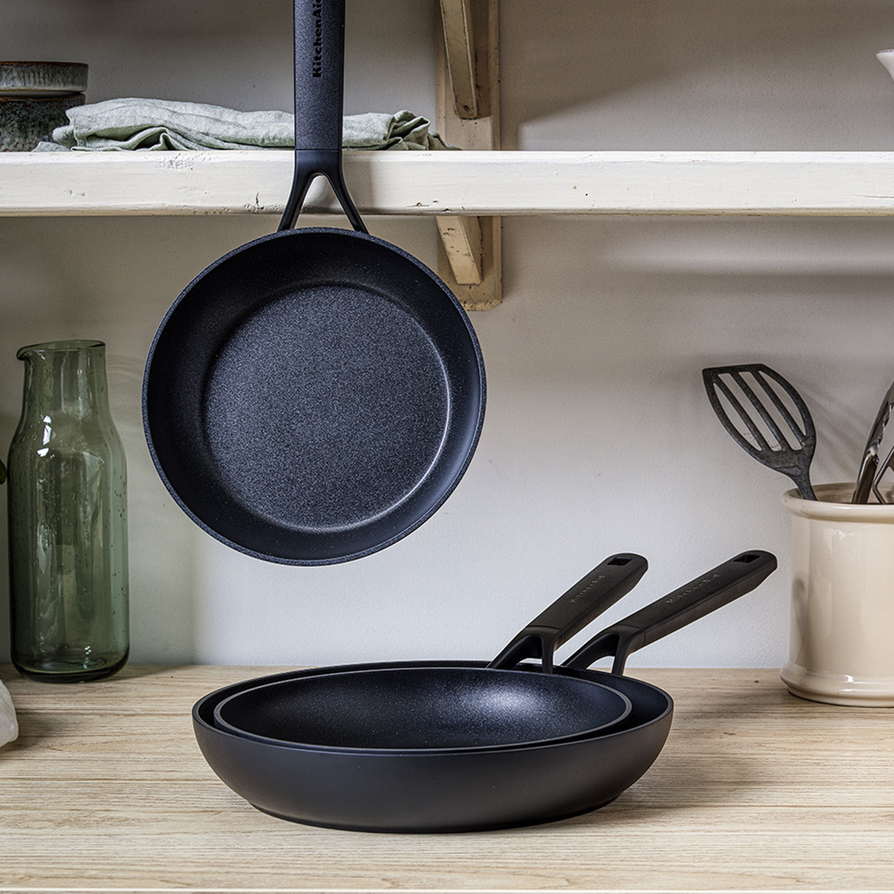Pfannen auf einer Holztheke. Eine Pfanne hängt, zwei andere sind gestapelt. Marke KitchenAid.