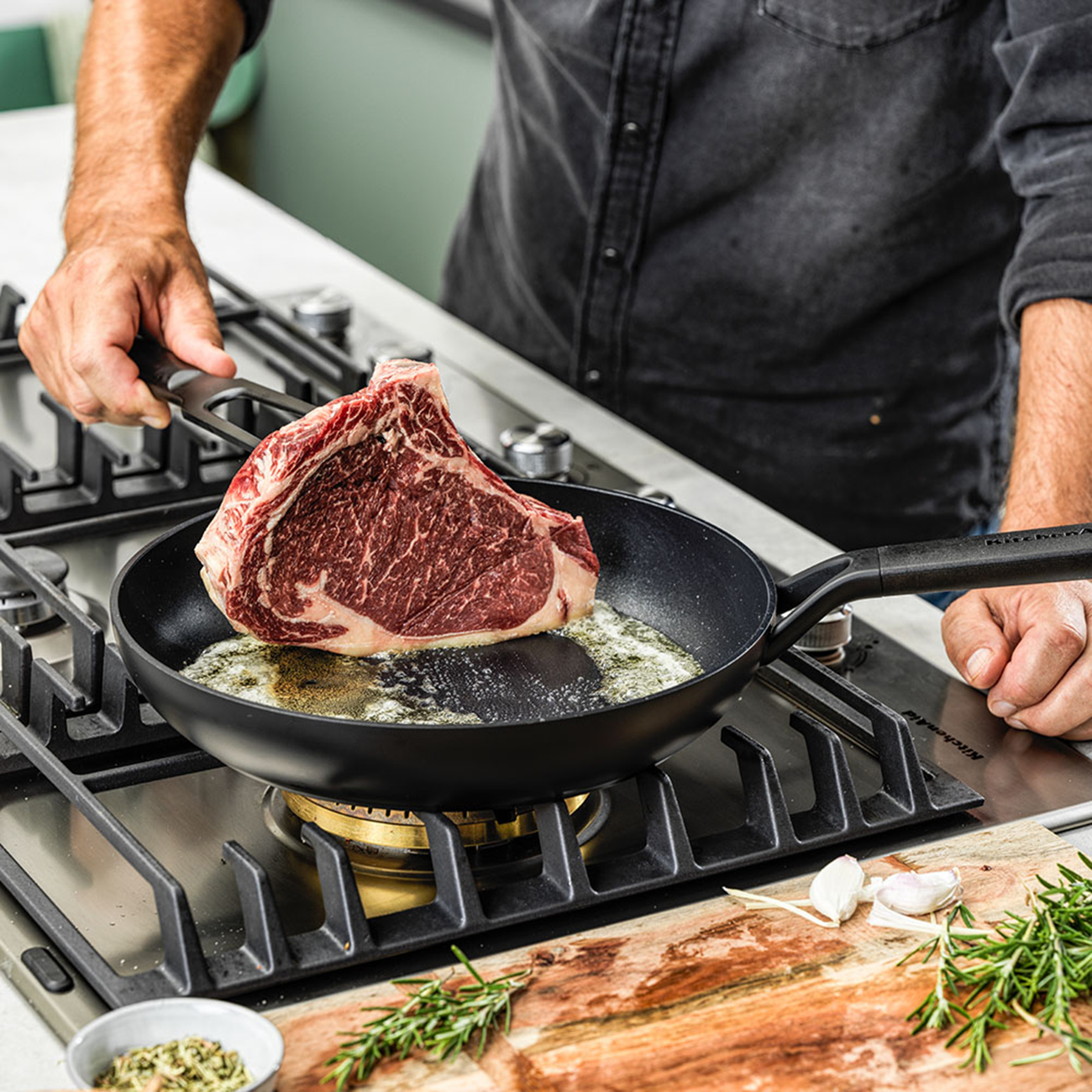 Eine Person brät ein großes Steak in einer Bratpfanne mit Butter auf einem Gasherd. Rosmarin und Knoblauch sind in der Nähe.