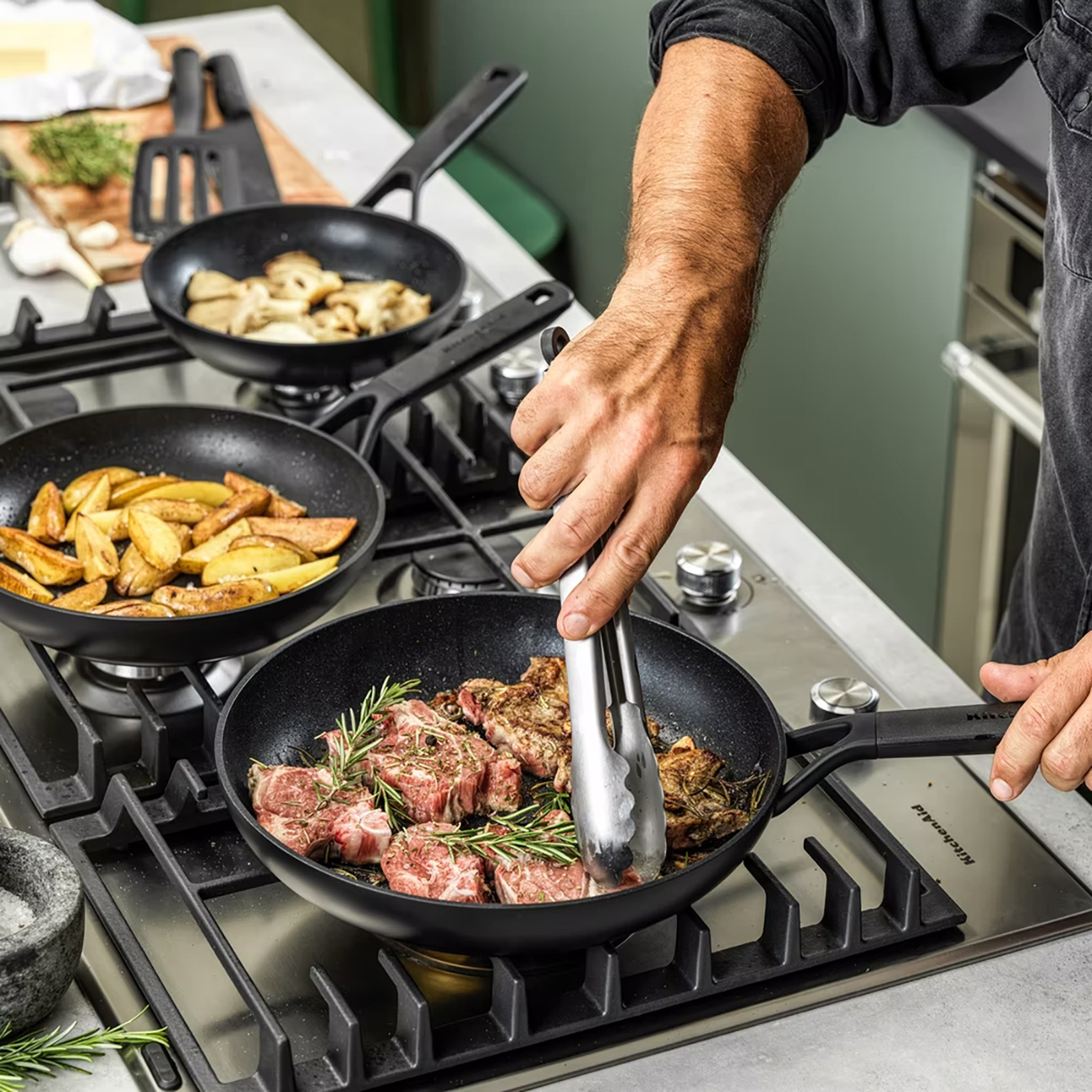 Koch wendet Fleisch mit Zangen in einer Pfanne. Weitere Pfannen mit Essen stehen auf dem Herd.