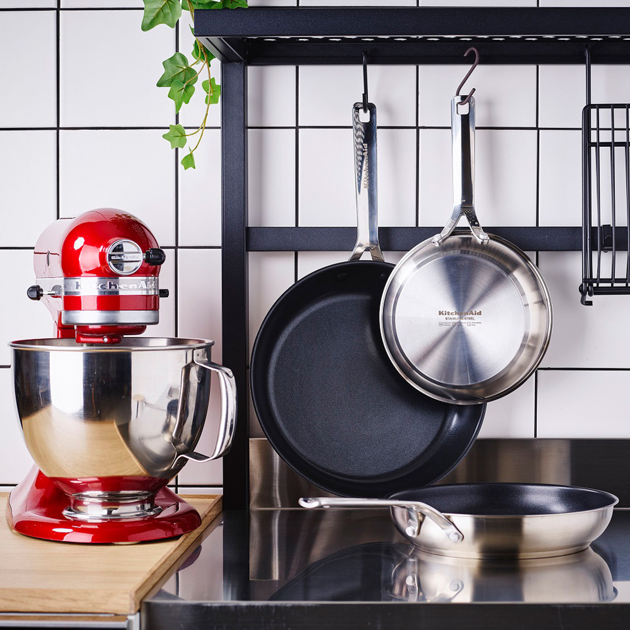 KitchenAid-Küchenmaschine, Pfannen und ein Gestell. Die Pfannen sind schwarz und silber. Weißer Fliesen-Hintergrund.