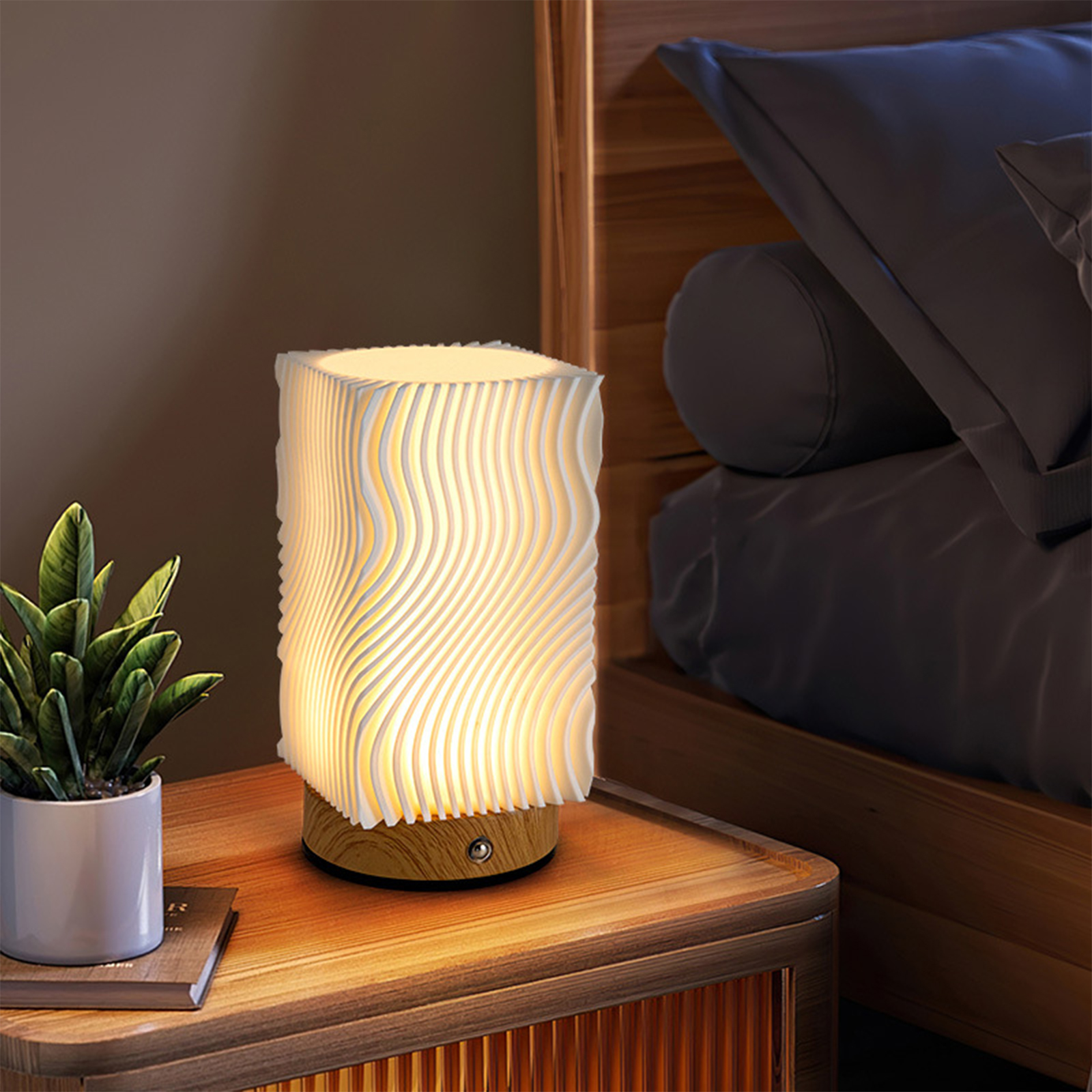 Weiße Lampe mit Wellenmuster auf Nachttisch. Kleine Pflanze und Buch sind daneben.