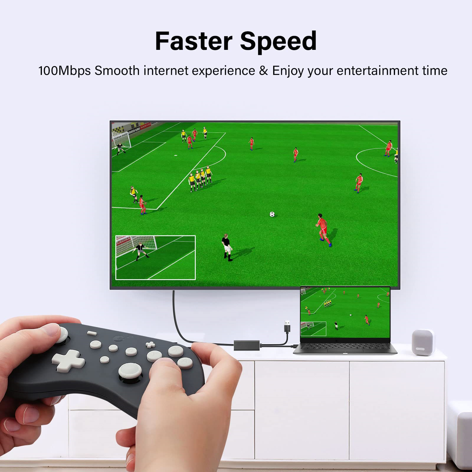 Eine Person, die einen Controller hält, spielt ein Fußballspiel auf einem Fernseher. Es gibt einen Laptop und einen Lautsprecher.