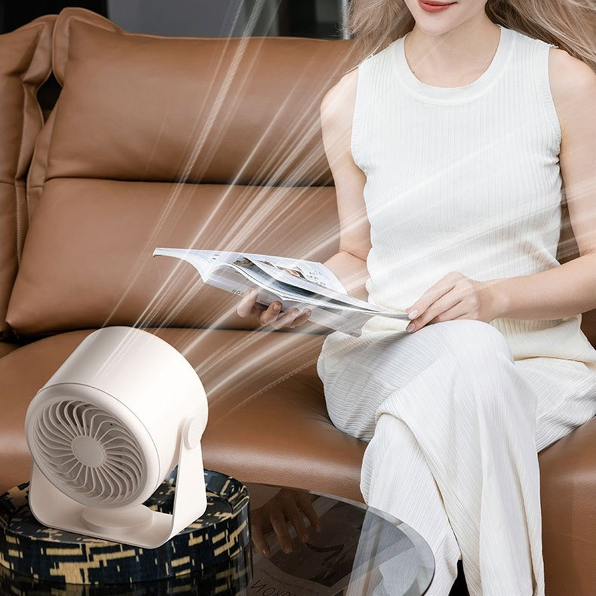 Frau sitzt auf einem braunen Sofa, hält eine Zeitschrift. Ein weißer Ventilator steht auf einem Tisch und bläst Luft.