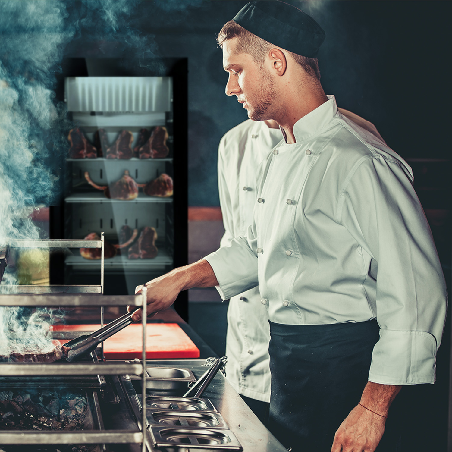 Due chef in cucina, uno che cucina una bistecca, un altro che guarda un'esposizione di carne sullo sfondo.