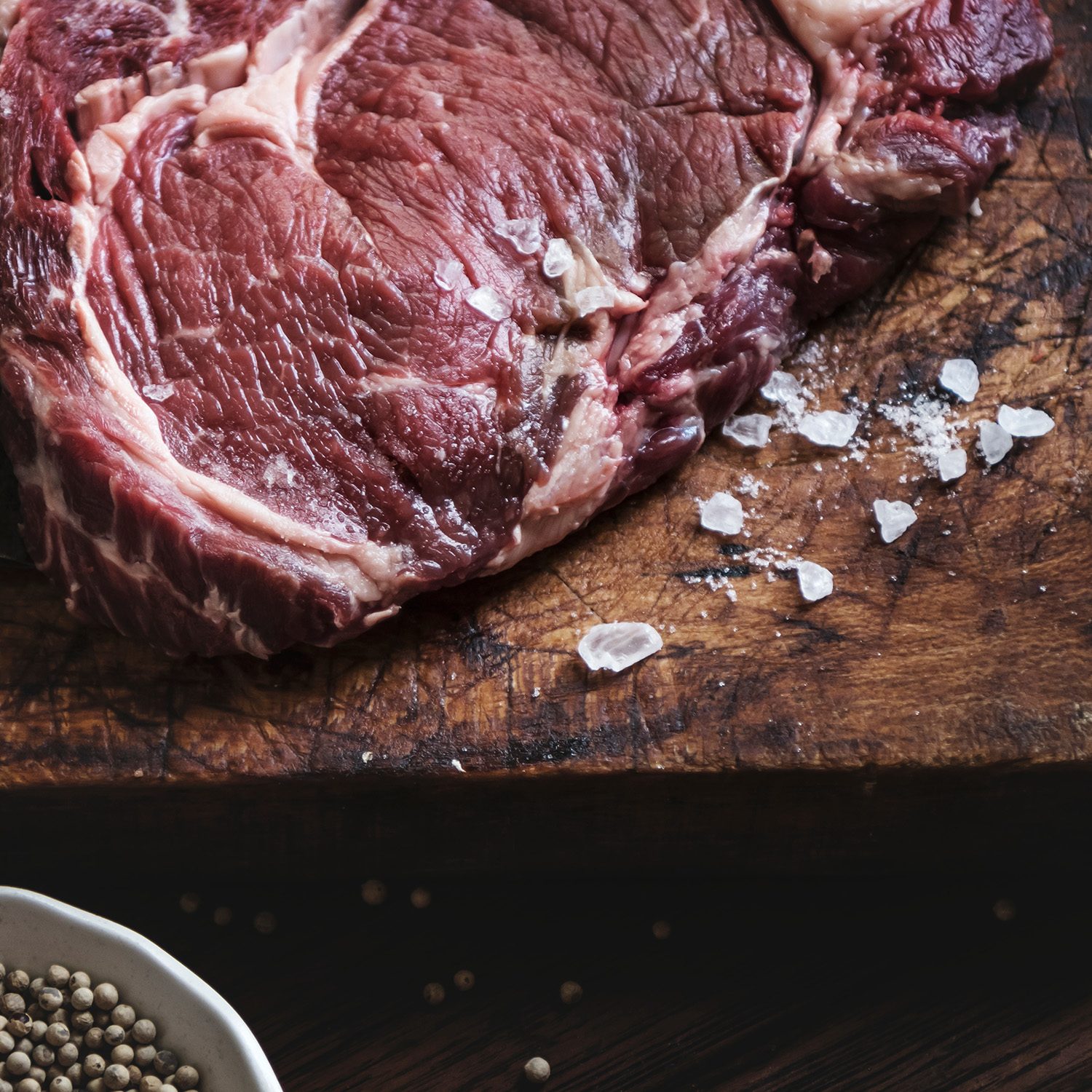 Una bistecca di ribeye cruda e rossa su un tagliere di legno con cristalli di sale sparsi.