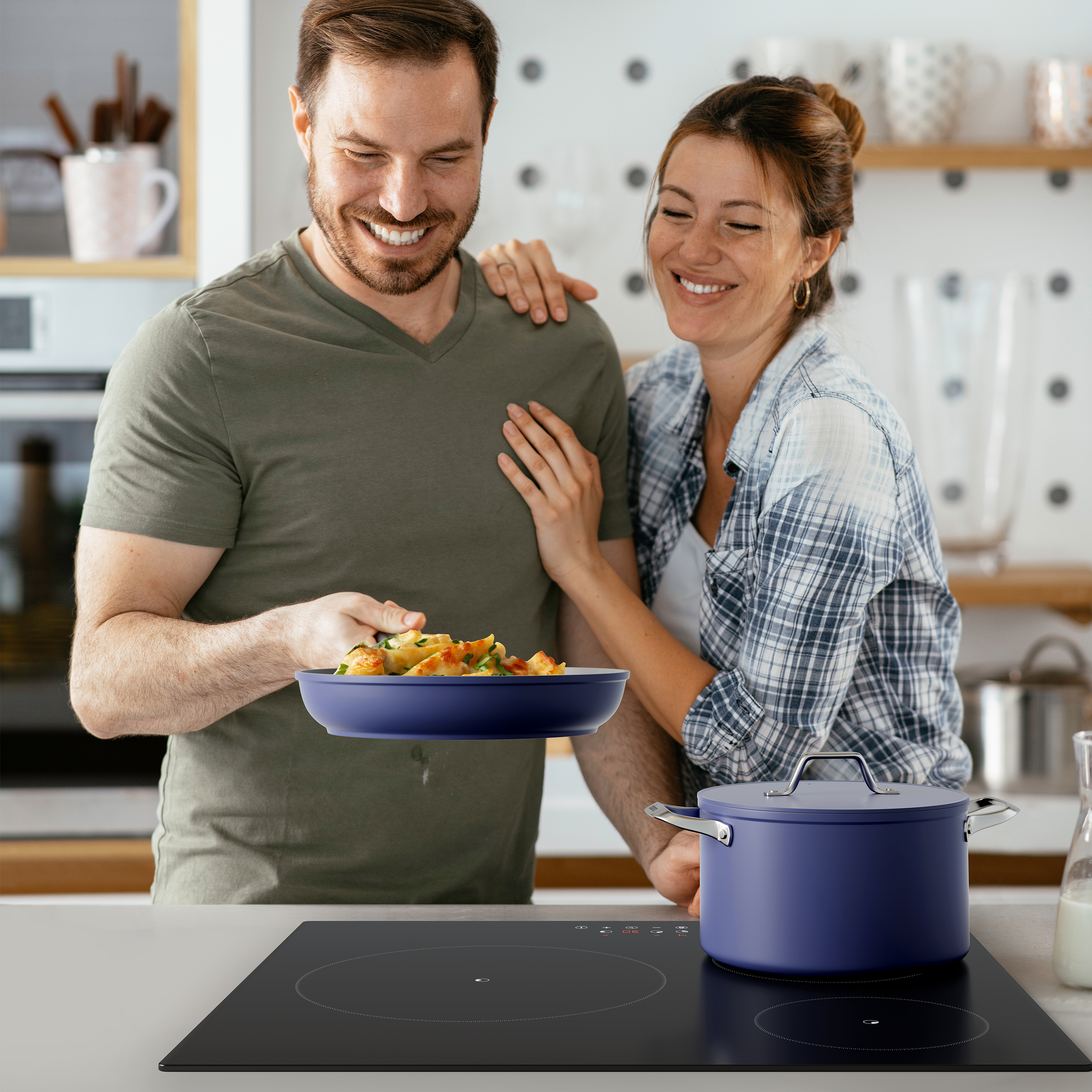 Un couple souriant tenant une assiette de nourriture au-dessus d'une plaque à induction avec une casserole bleue.