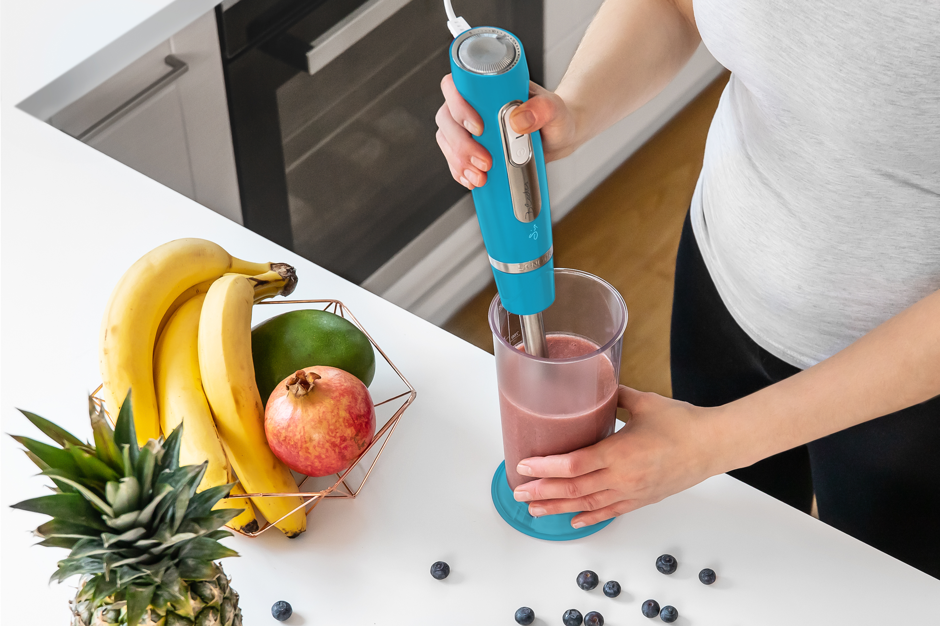 Kobieta miksuje smoothie niebieskim blenderem. Banany, mango, granat i jagody na blacie.