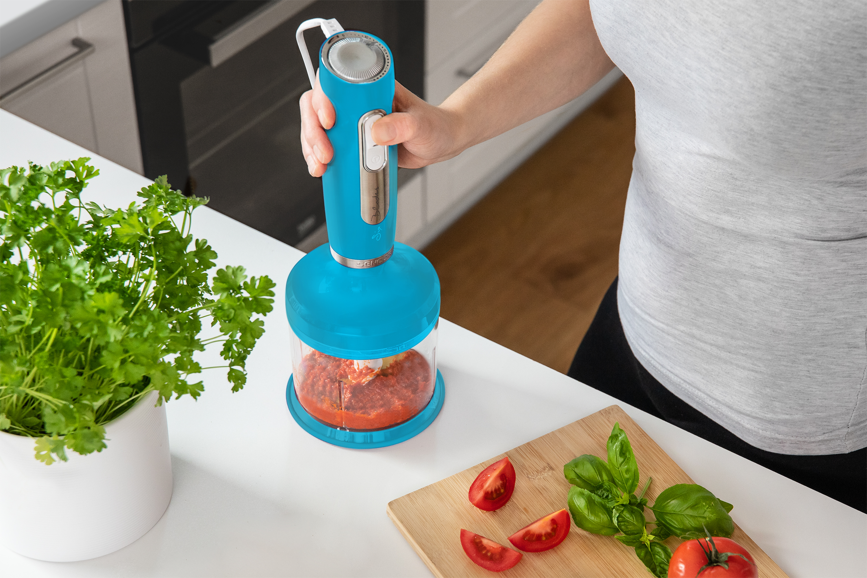 Kobieta używa niebieskiego blendera. Pomidory, bazylia i drewniana deska do krojenia na blacie.