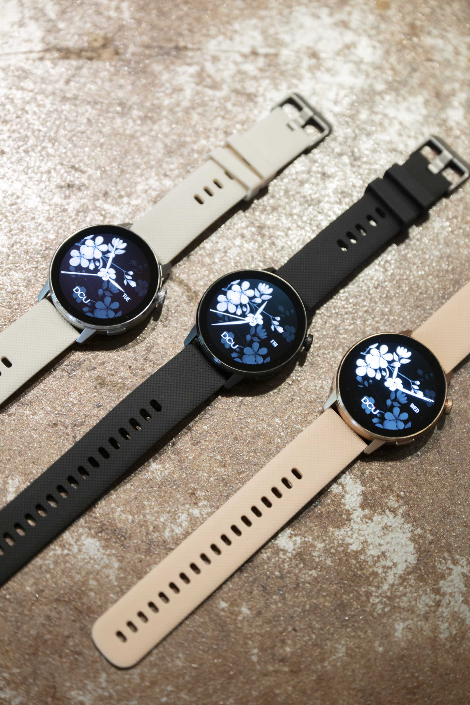 Trzy smartwatche z kwiatowymi wyświetlaczami na ekranach, z paskami w różnych kolorach na teksturowanej powierzchni.