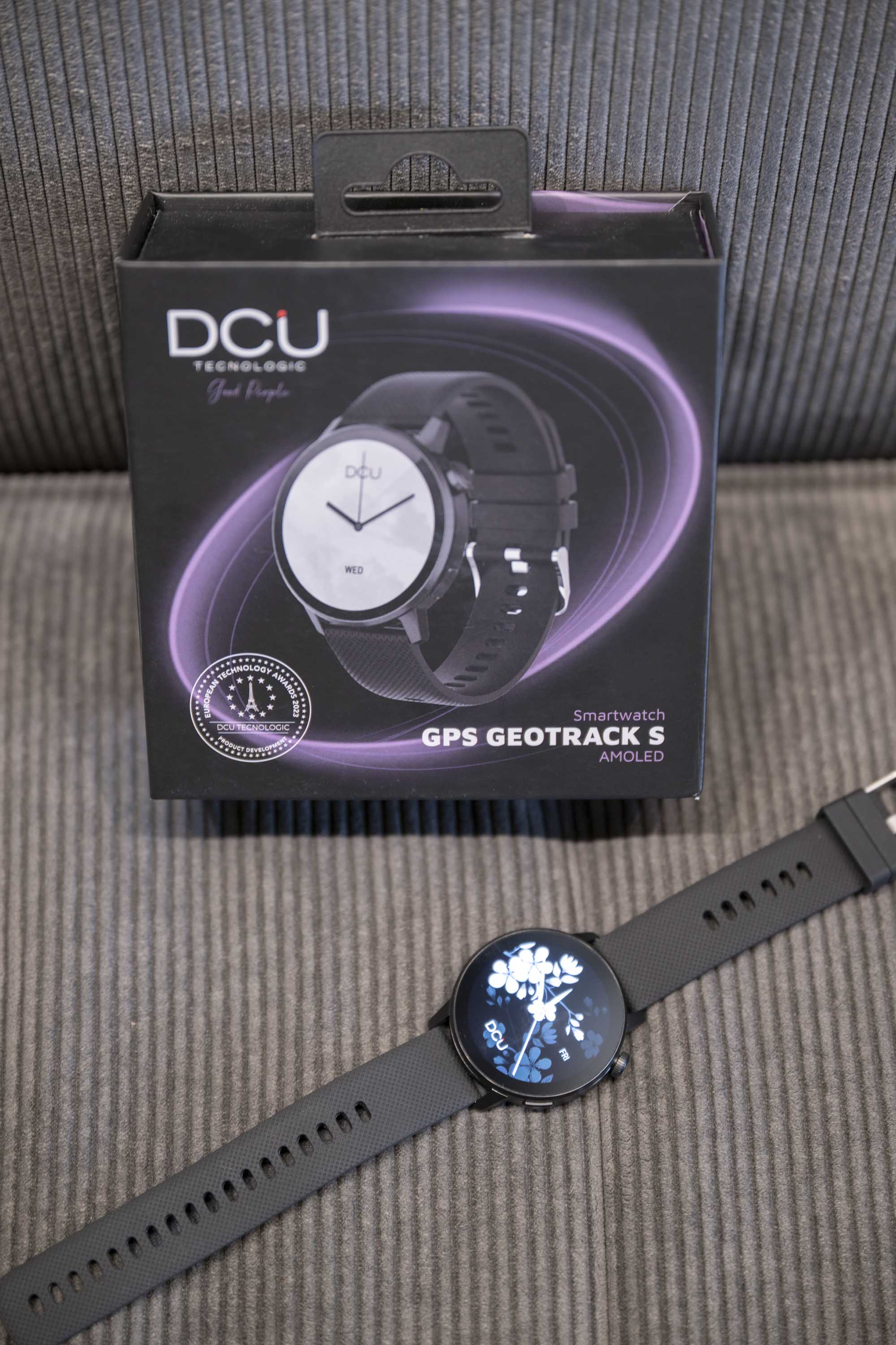 Czarny smartwatch z czarnym paskiem i pudełkiem. Pudełko ma obraz zegarka i tekst.