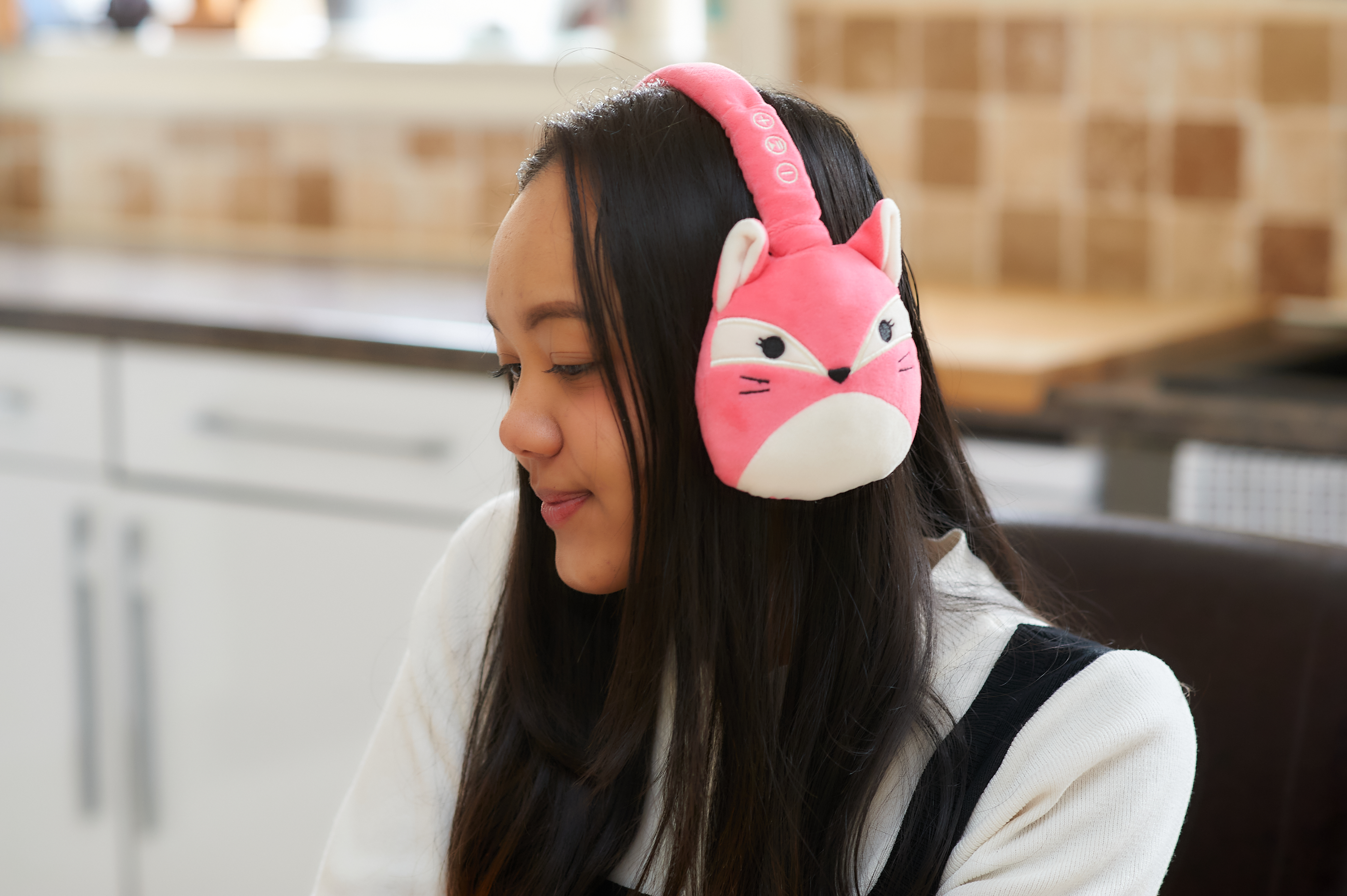 Een jonge vrouw met roze vossenoor-koptelefoon, in een thuisomgeving, glimlachend.