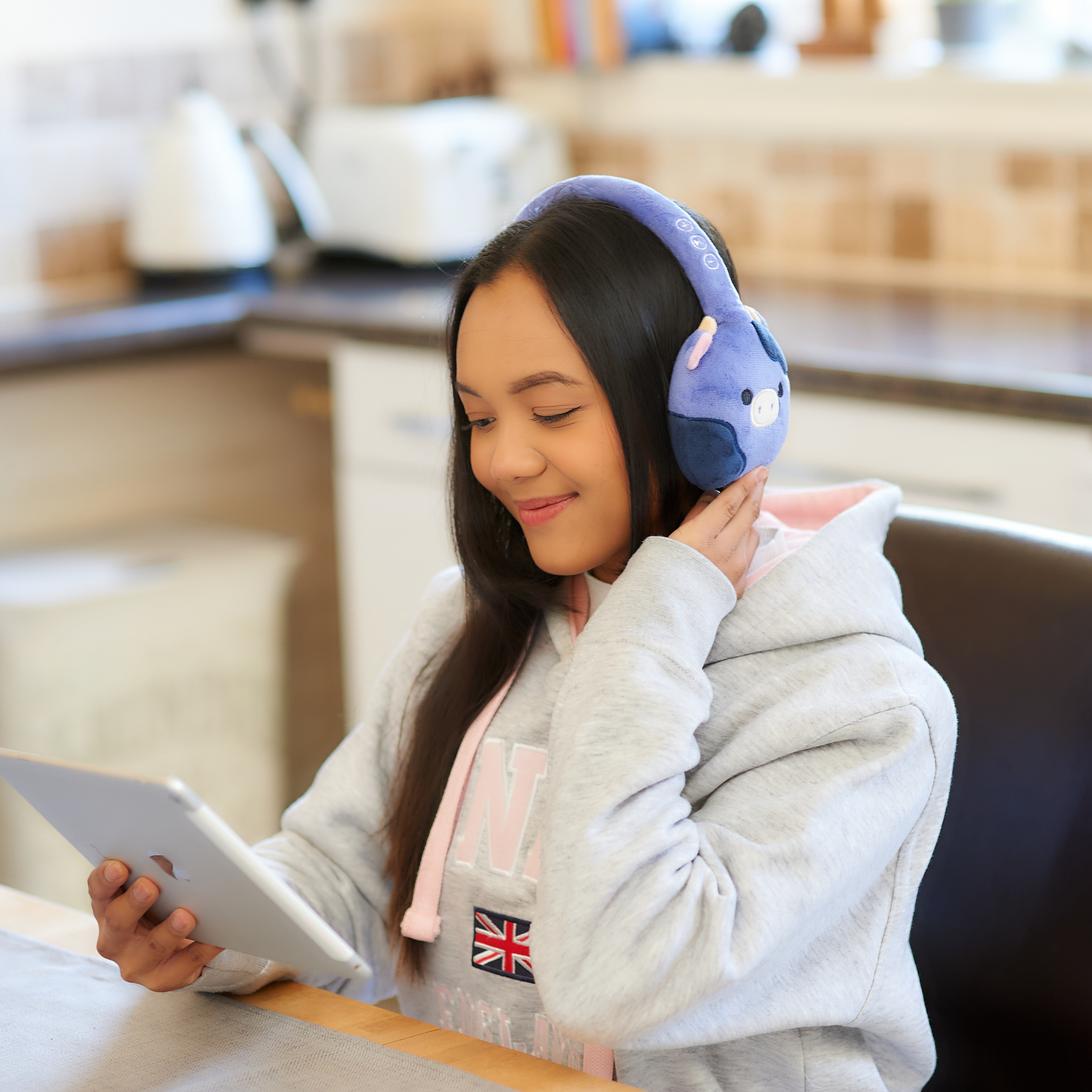 Jonge vrouw met een blauwe koptelefoon, glimlachend terwijl ze een tablet vasthoudt. Keukenachtergrond.