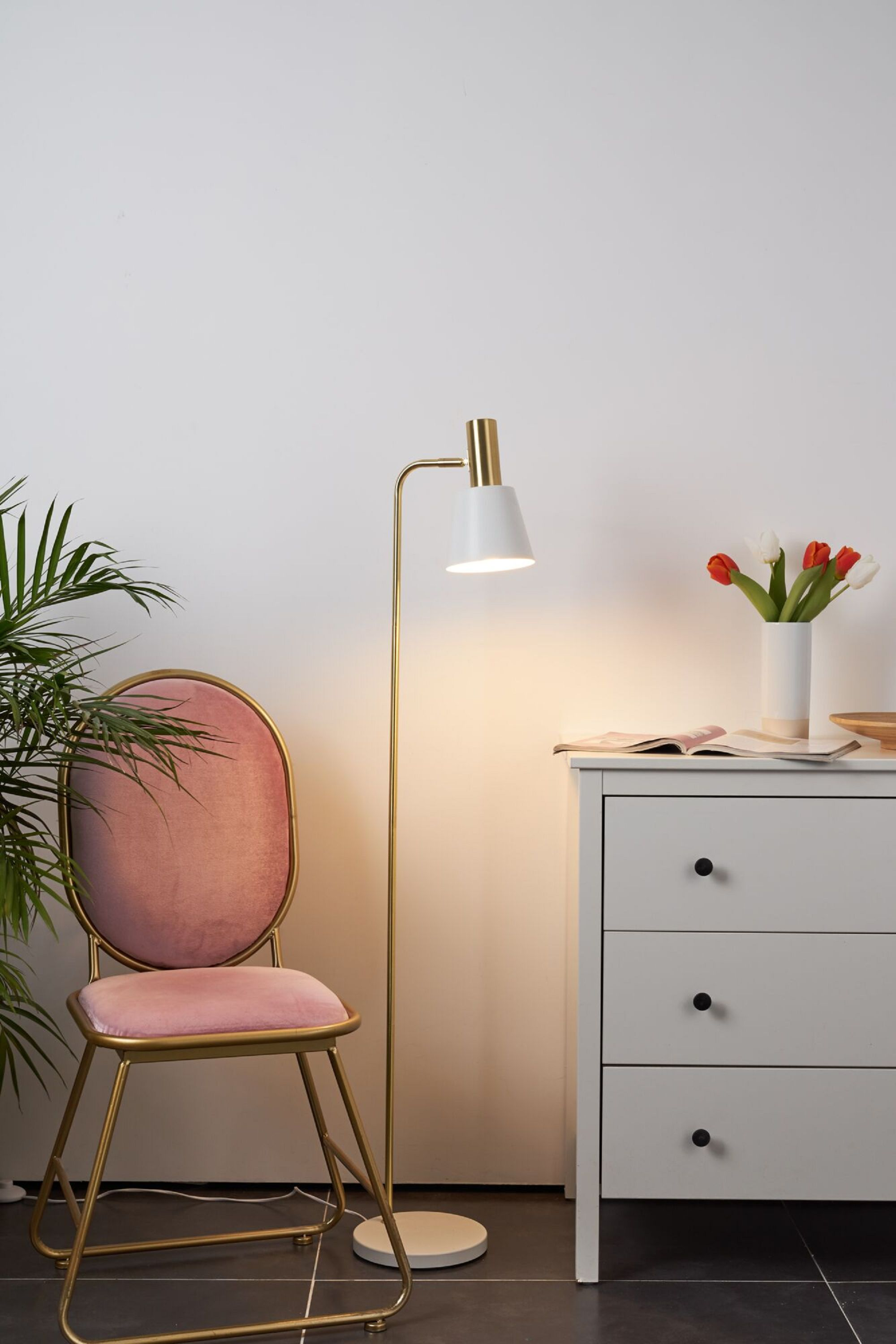 Eine Stehlampe, ein rosa Stuhl und ein weißer Schrank mit Blumen stehen vor einer weißen Wand.