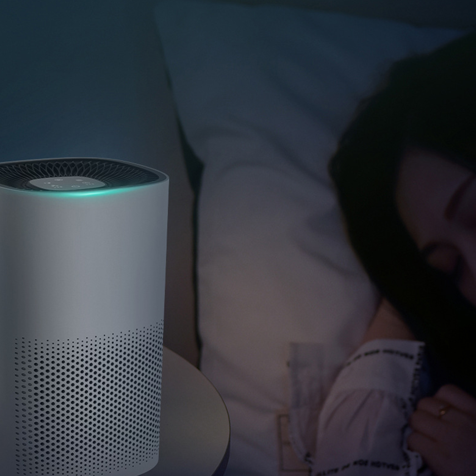 Ein weißer Luftreiniger auf einem Tisch neben einem Bett. Eine Frau schläft im Bett. Der Raum ist schwach beleuchtet.
