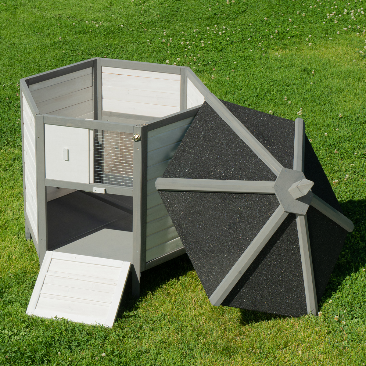 Ein hölzerner Hühnerstall mit offenem Dach und Rampe, auf einer grünen Wiese. Der Stall ist weiß und grau gestrichen.