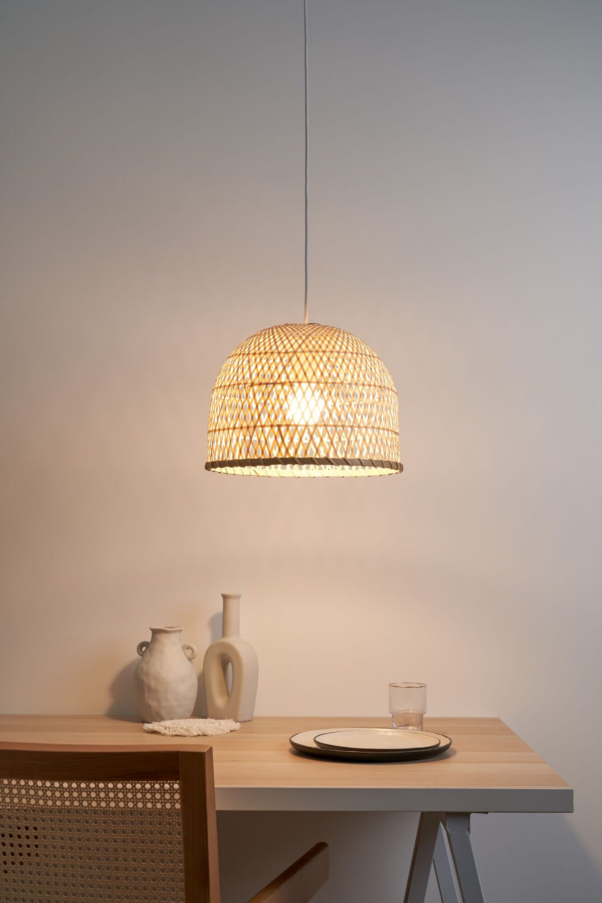 Wisząca lampa z wikliny nad drewnianym stołem z talerzem, szklanką i wazonem.