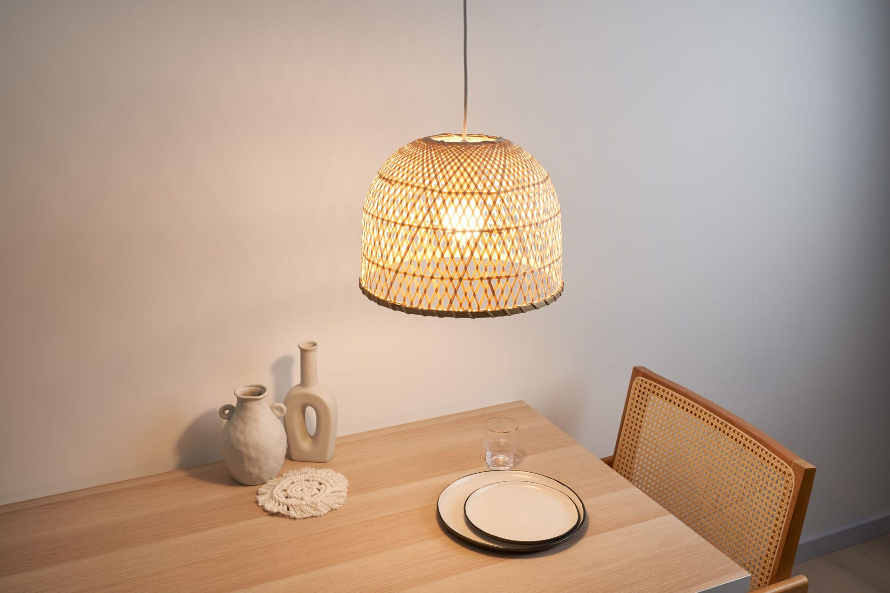 Tkana lampa wisząca z wikliny wisząca nad drewnianym stołem. Są wazony i talerze.