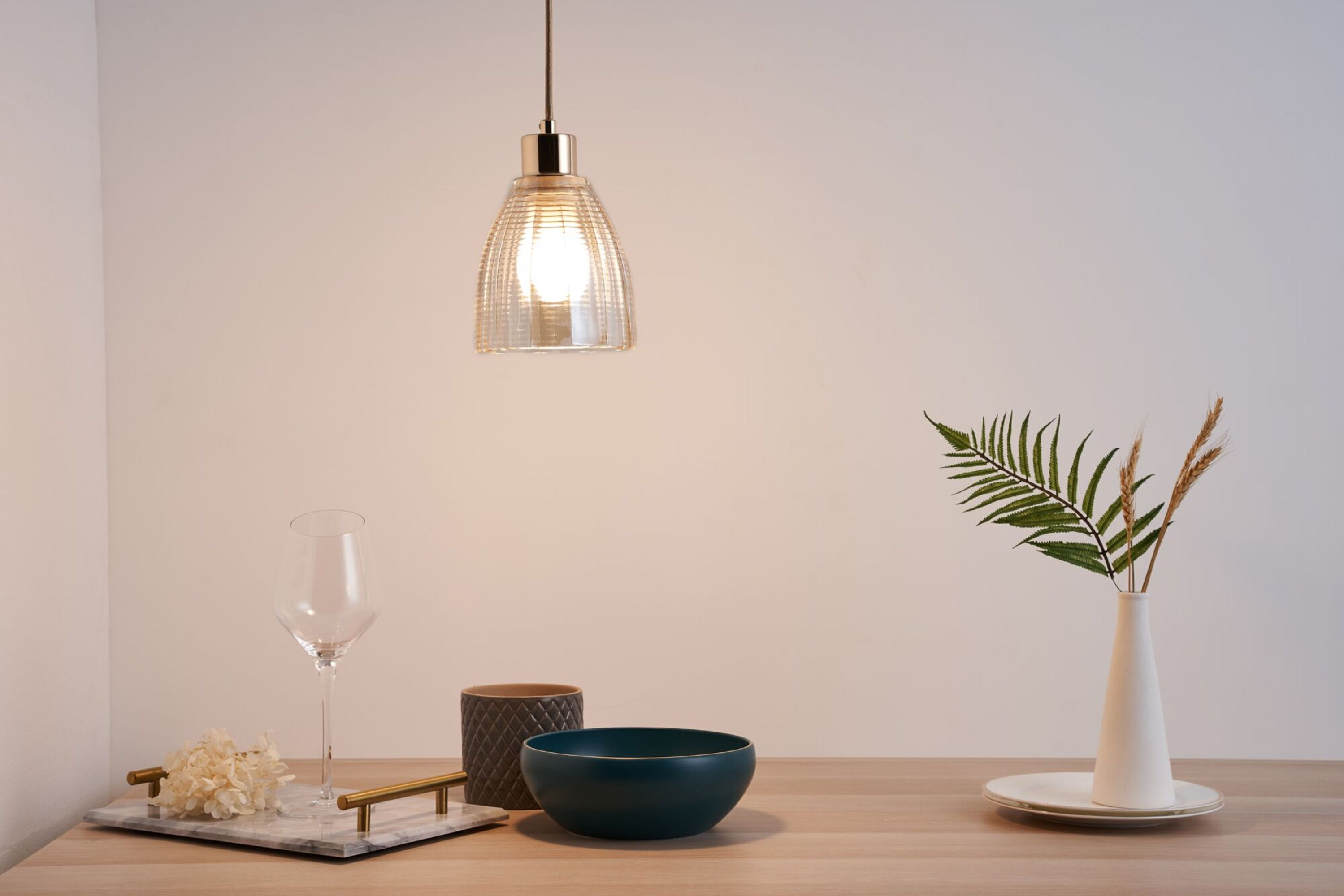 Lampa wisząca nad stołem. Szkło, miska i wazon z paprocią i pszenicą. Jasne tło.