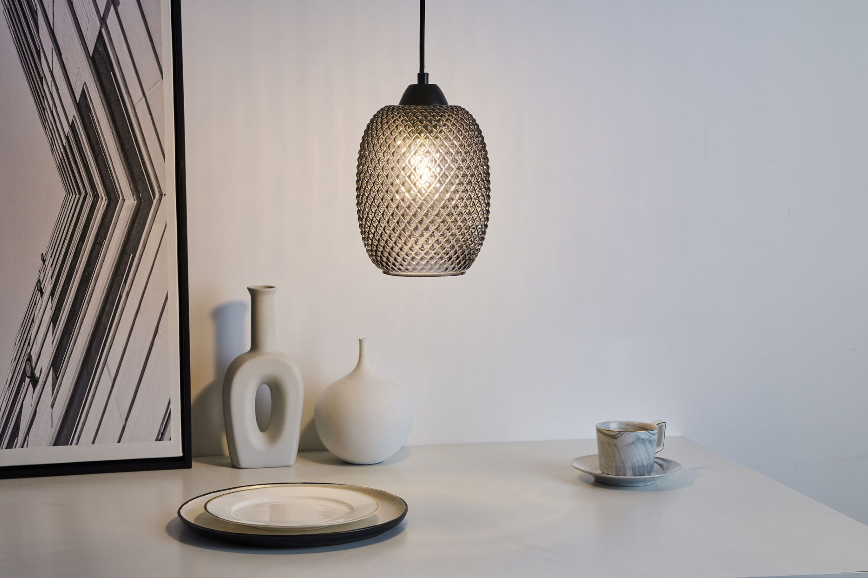 Hängeleuchte mit strukturiertem Glasschirm. Weißer Tisch, Vasen und eine Tasse.