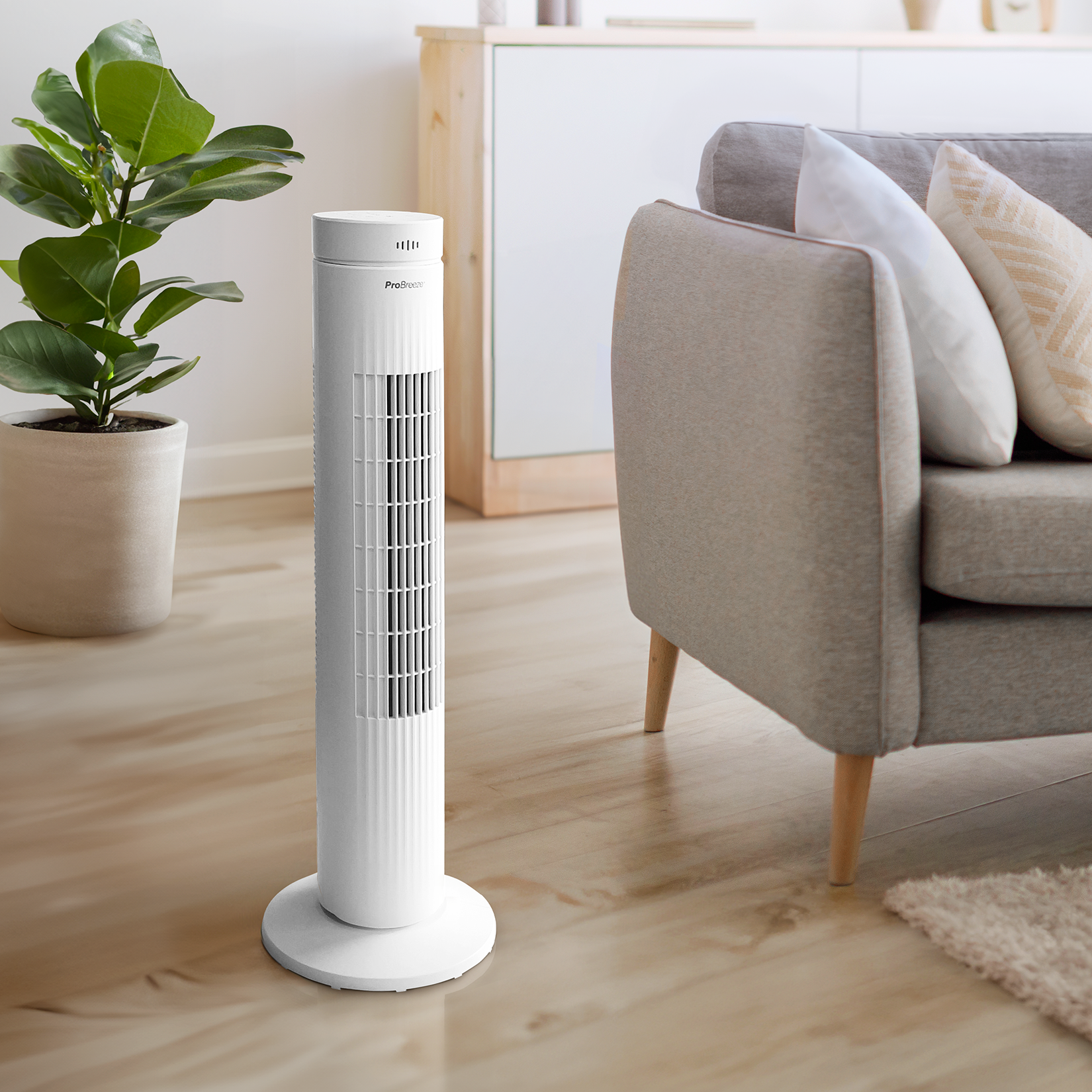 Weißer Turmventilator auf Holzboden, mit Sofa und Pflanze im Hintergrund. Innenaufnahme.