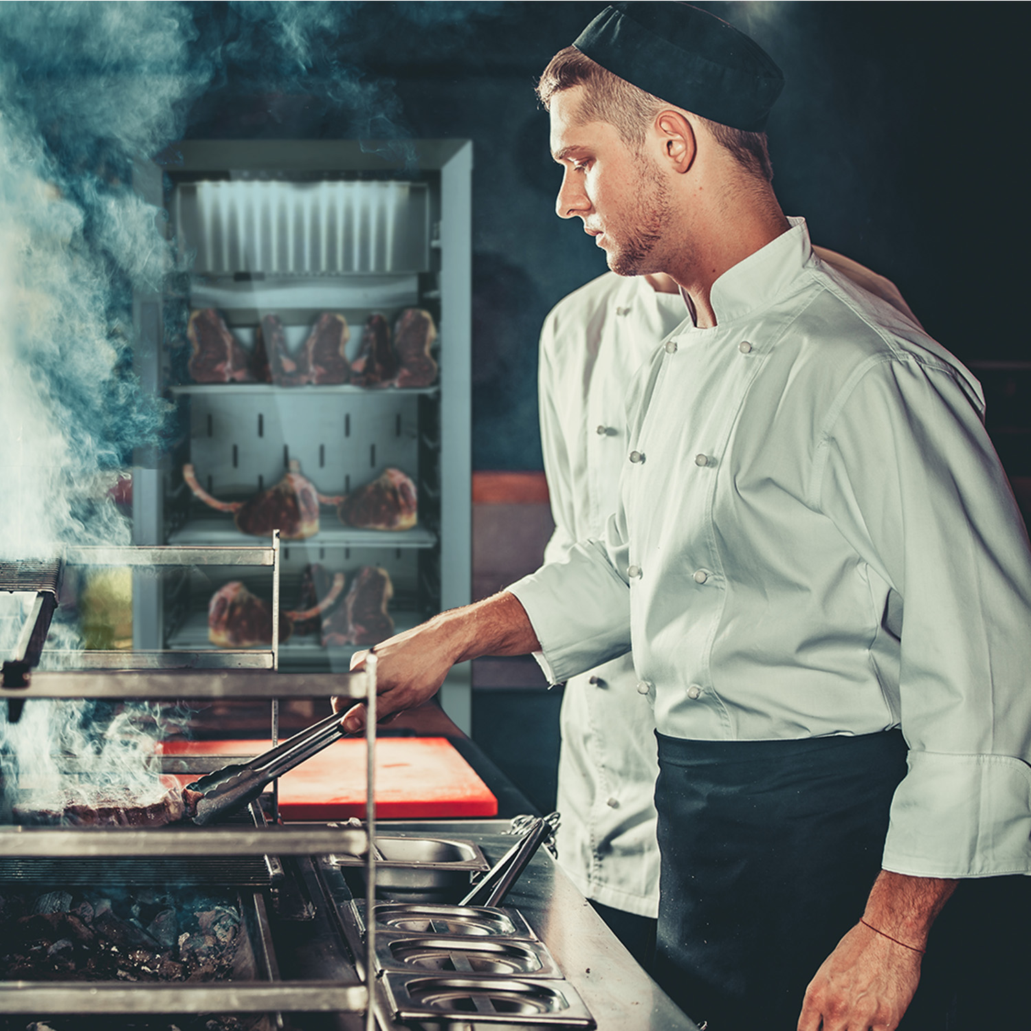 Ein Koch grillt Fleisch mit einer Zange. Im Hintergrund befindet sich ein Kühlschrank mit Fleisch.