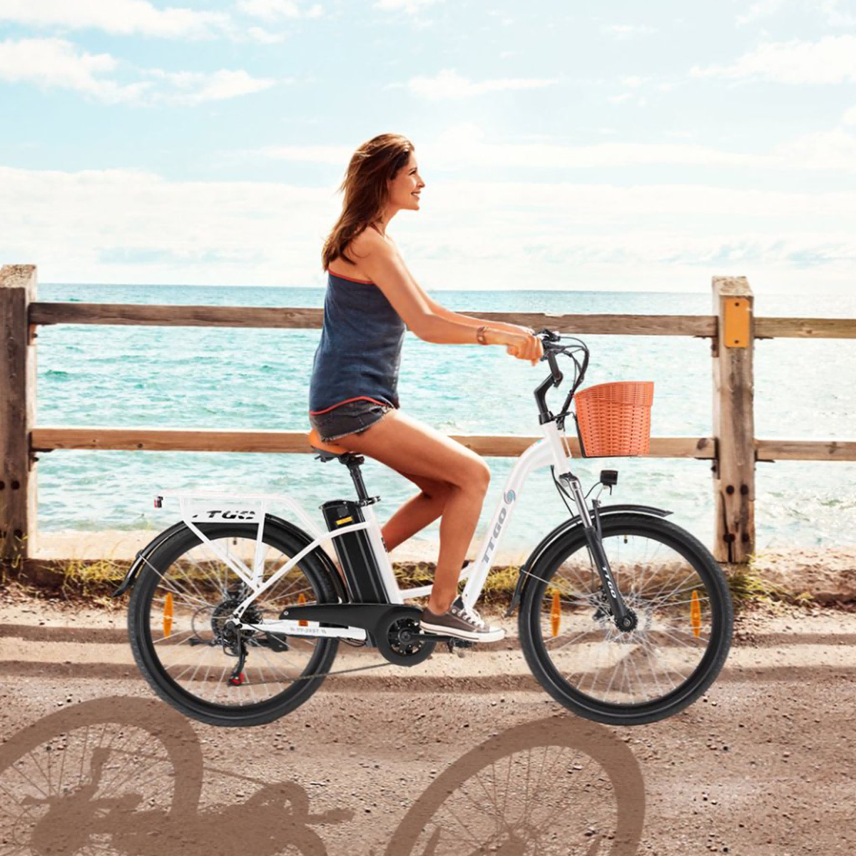 Kobieta jedzie na białym rowerze elektrycznym na ścieżce na plaży. Koszyk z przodu. Morze i niebo w tle.