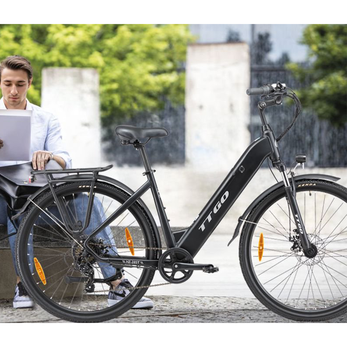 Mężczyzna czyta siedząc obok czarnego roweru elektrycznego TTGO w słoneczny dzień, w otoczeniu miejskim.