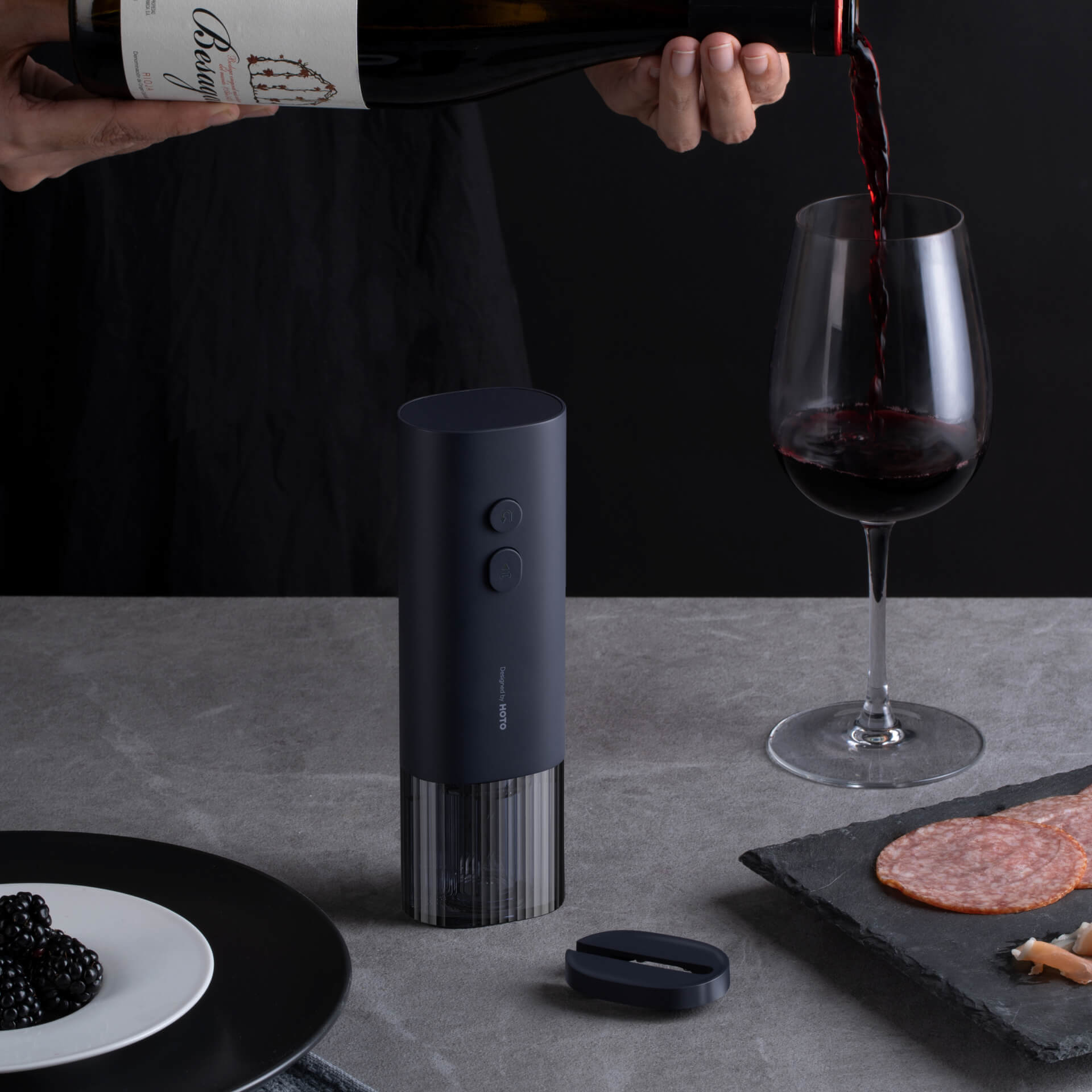 Eine Hand gießt Rotwein in ein Glas. Ein Weinöffner und andere Lebensmittel sind auf dem Tisch.