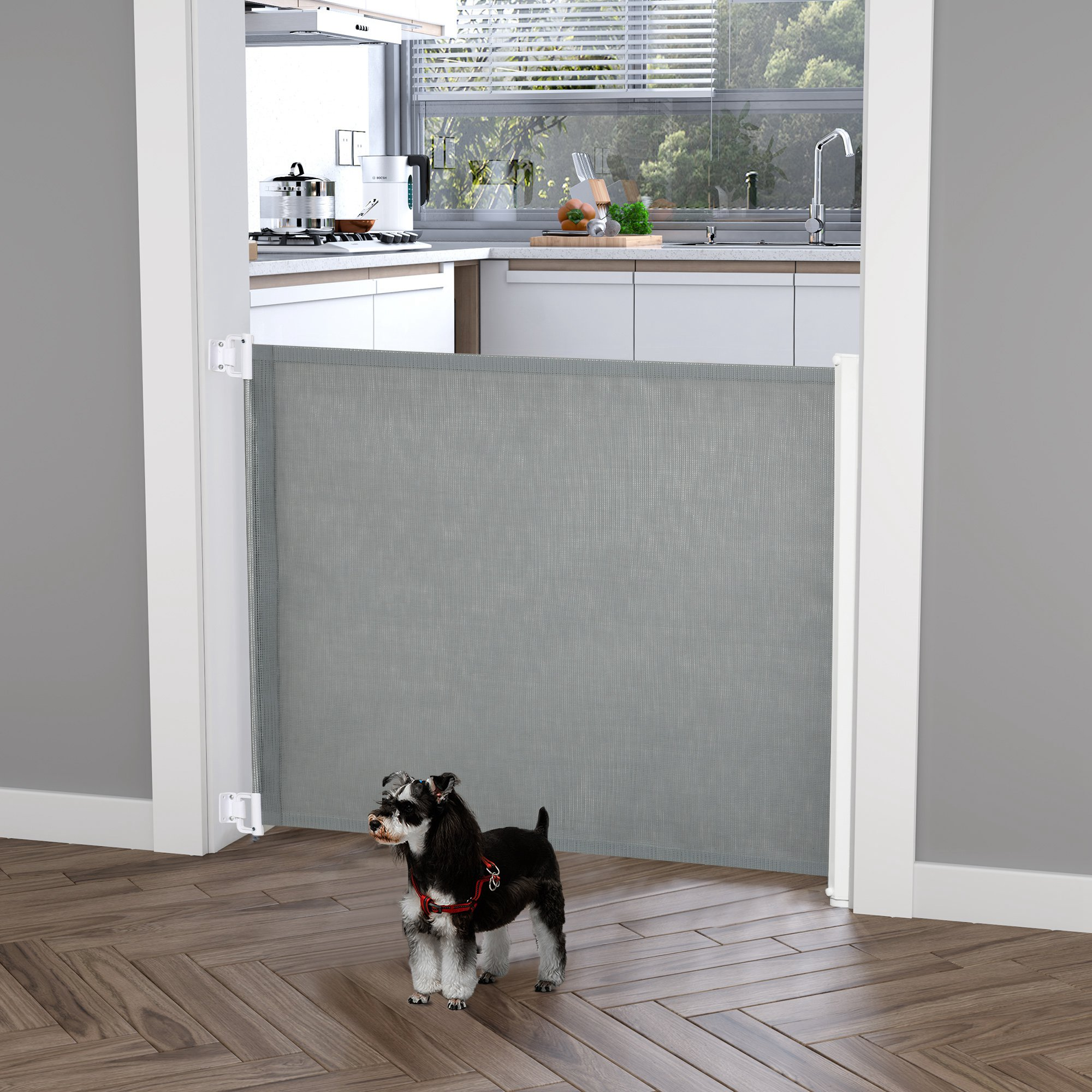 Un perro pequeño está en una puerta bloqueada por una barrera retráctil gris para mascotas, a una cocina.