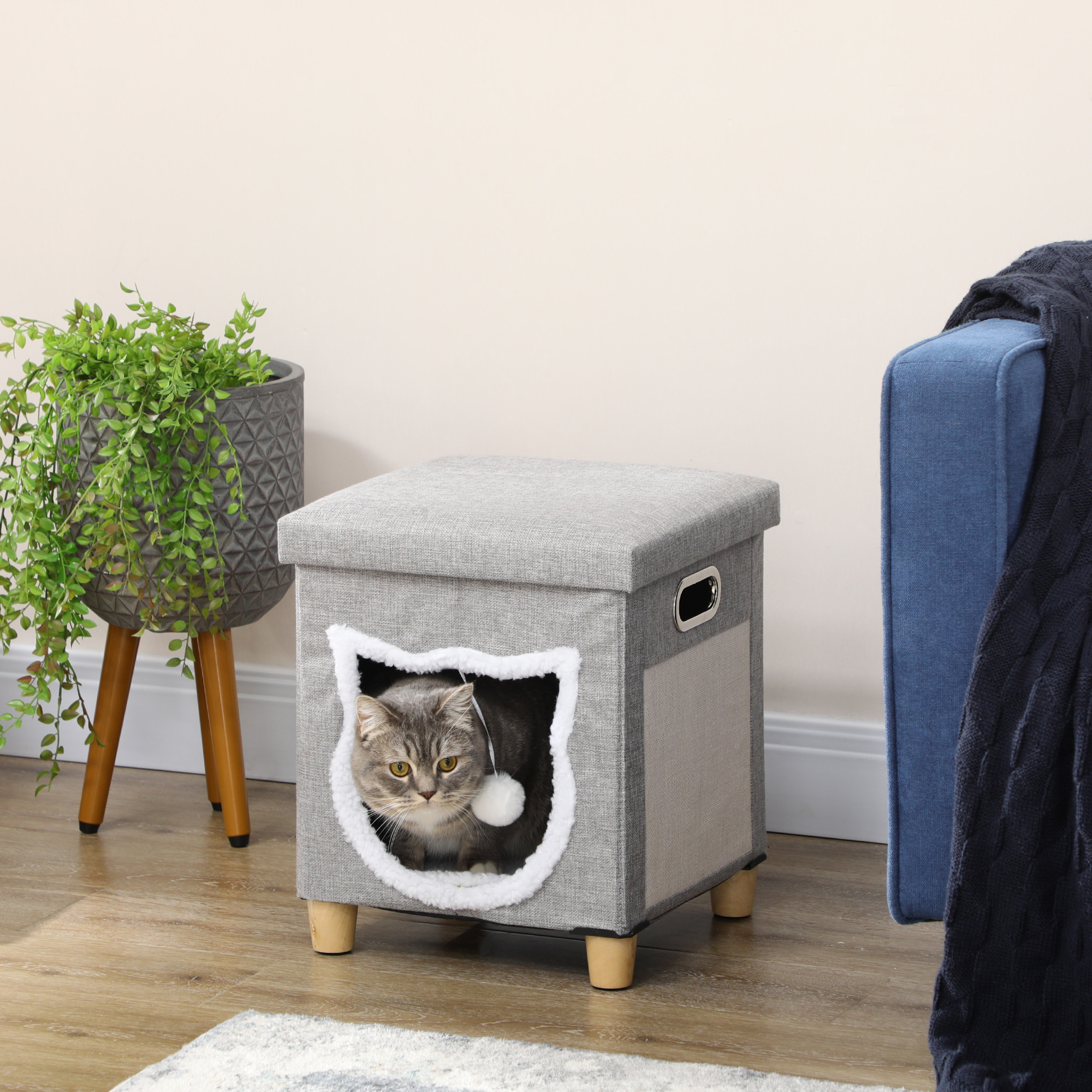 Una cama gris para gatos con un gato dentro. La cama está en un piso de madera, cerca de una planta y un sofá azul.
