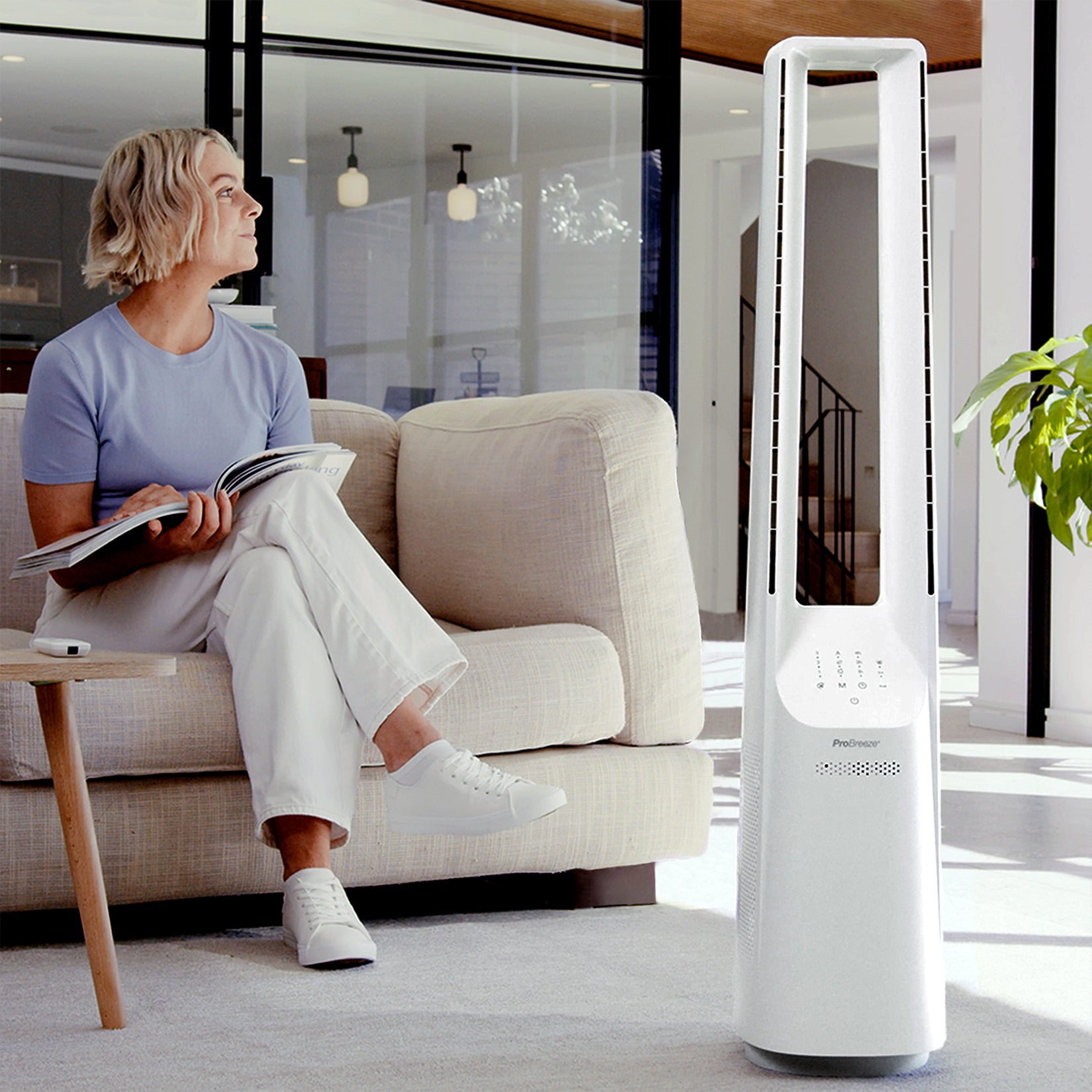 Frau liest in der Nähe eines weißen Ventilators, beiges Sofa und Fenster, heller Raum.