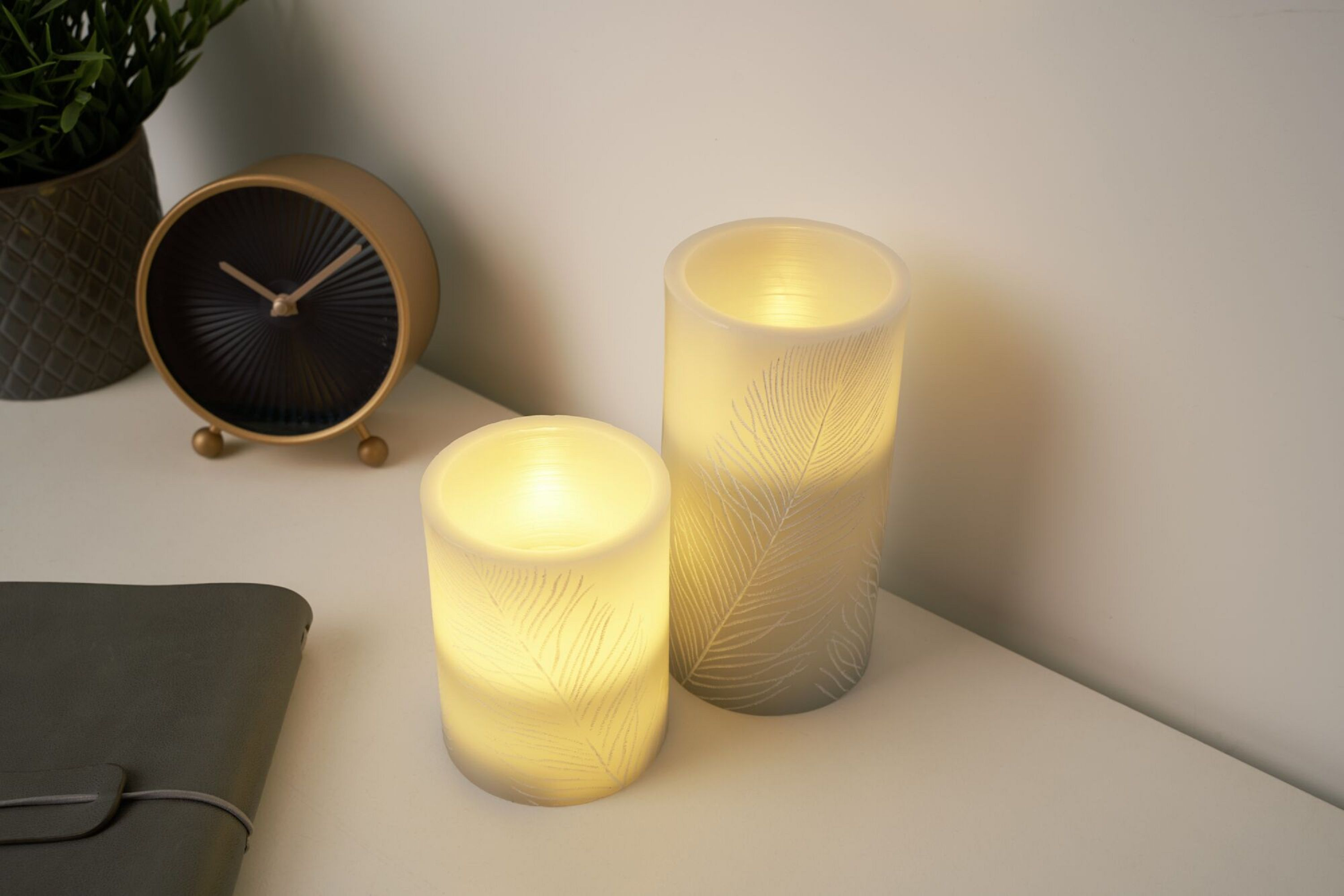 Dwa świecące światła w kształcie świec ze wzorem piór, zegar i roślina na białym stole.