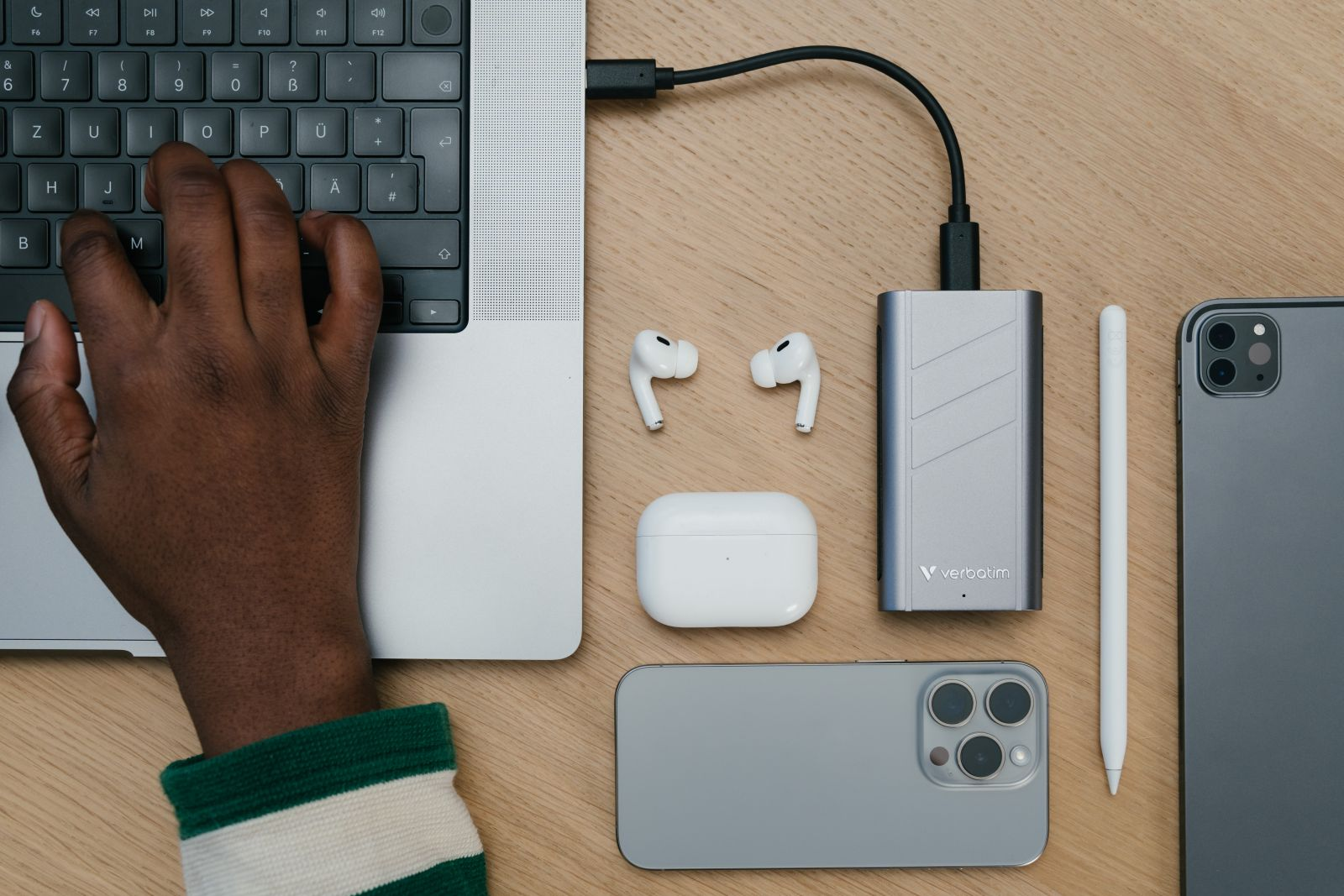 Ręka osoby na klawiaturze laptopa, z akcesoriami takimi jak AirPods, ładowarka i tablet.