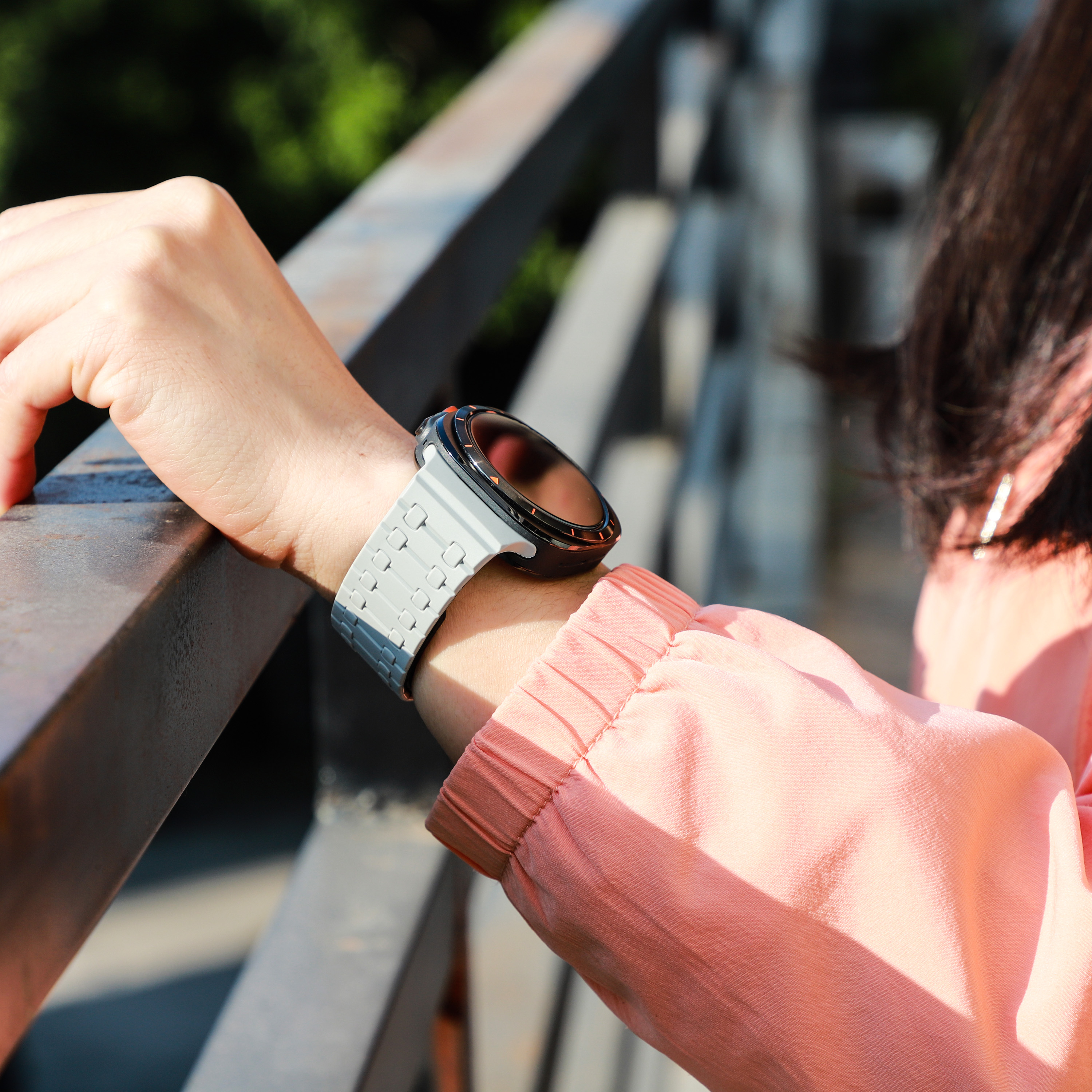 Das Handgelenk einer Person mit einer Smartwatch. Die Uhr hat ein graues Armband. Die Person ist draußen an einem Metallgeländer.