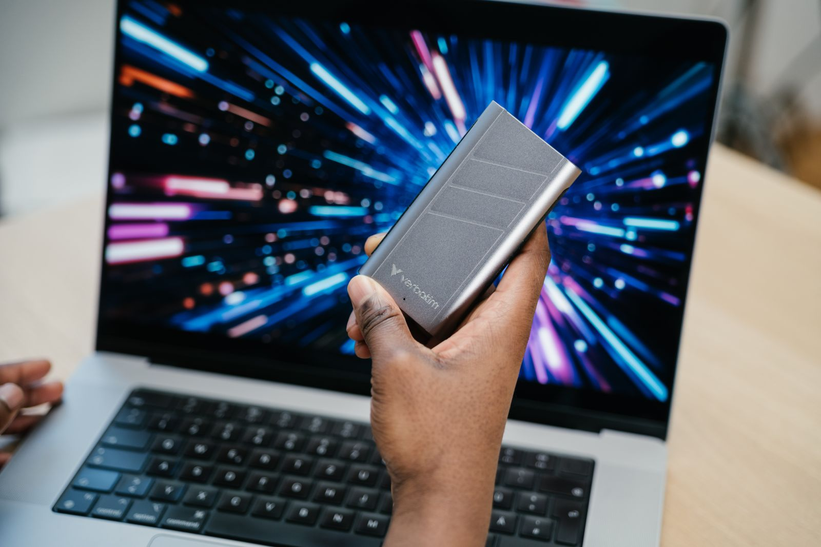 Ręka trzymająca szary zewnętrzny dysk twardy Verbatim przed laptopem z niebieskim tłem.
