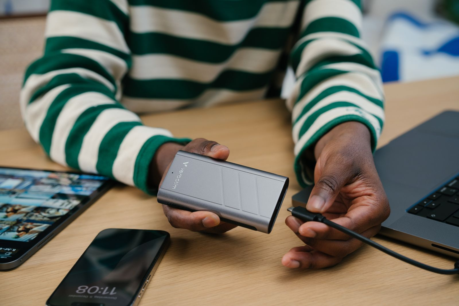 Osoba trzyma srebrny zewnętrzny dysk twardy. Laptop i telefon leżą na stole.