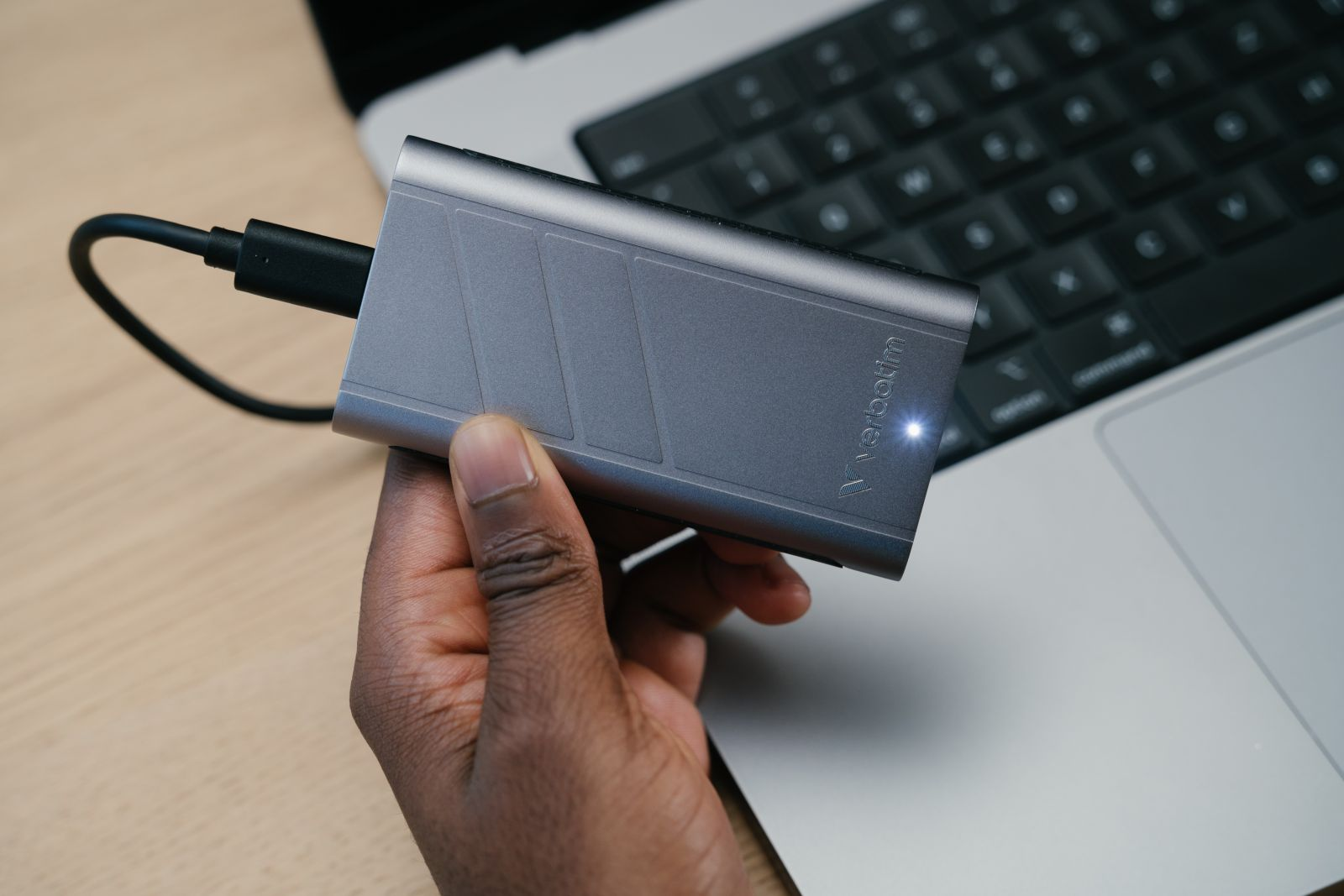 Dłoń trzymająca srebrny zewnętrzny dysk twardy podłączony do laptopa. Klawiatura laptopa jest widoczna.