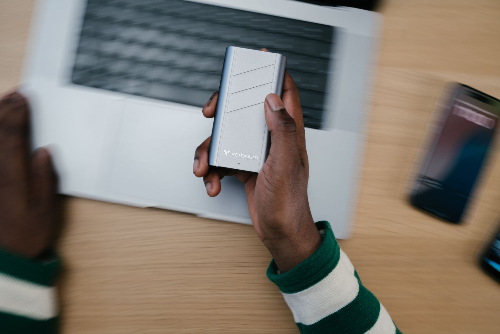 Ręka osoby trzymająca srebrny przenośny dysk twardy na biurku z laptopem i telefonem.