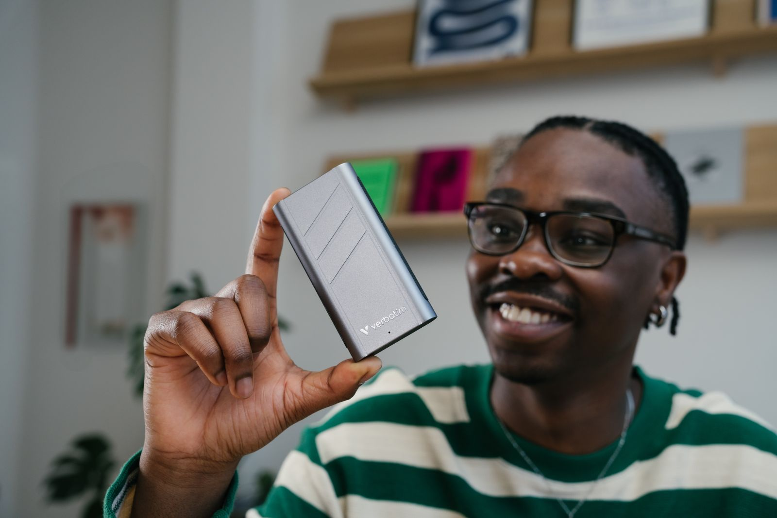 Mężczyzna w okularach, uśmiechnięty i trzymający szary prostokąt. Jest w pokoju z regałem na książki.