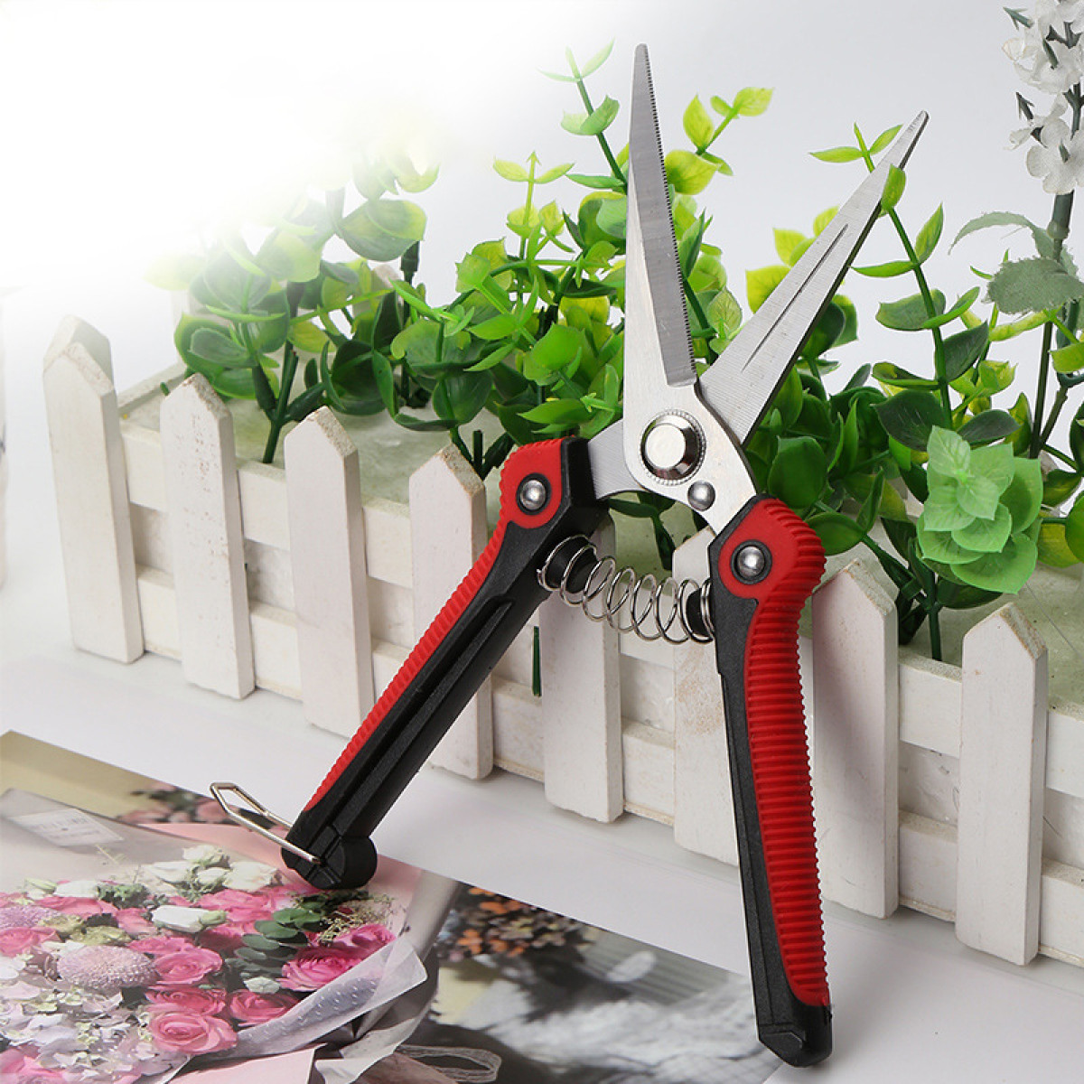 Rot-schwarze Gartenschere mit silberner Feder und Klingen, vor einem Blumendisplay.