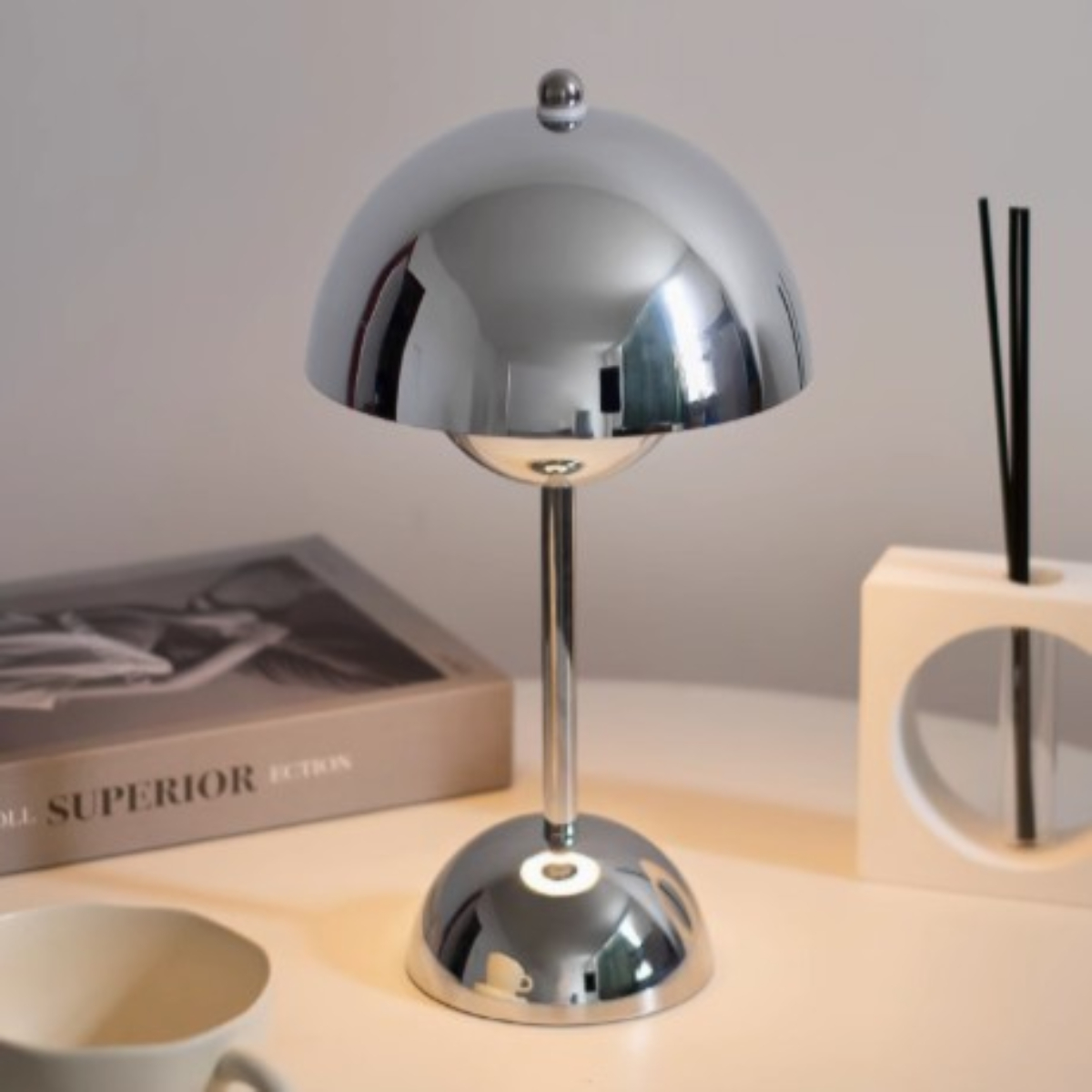 Silberne, kuppelförmige Schreibtischlampe. Sie steht auf einer weißen Oberfläche mit einem Buch und einem Holzdiffusor.
