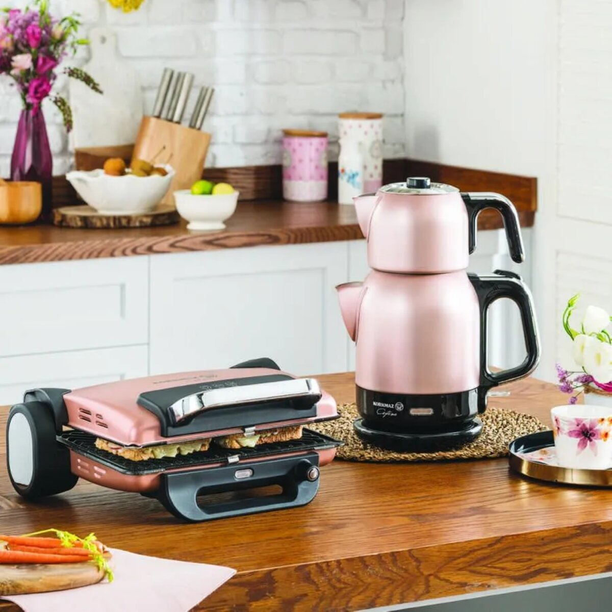 Pembe ve siyah çay makinesi ve sandviç makinesi seti, çiçekler ve sebzelerle birlikte ahşap bir mutfak tezgahında.
