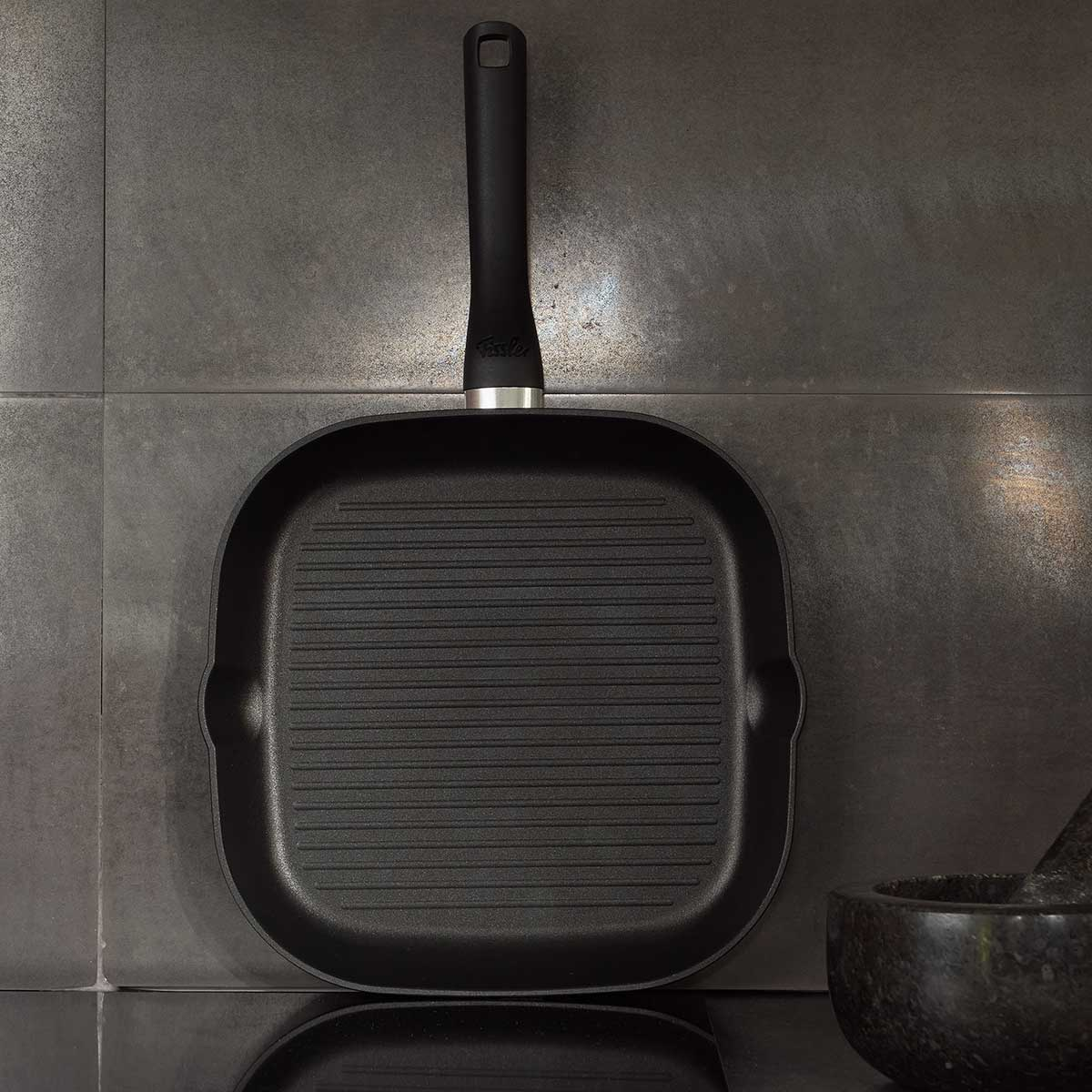 Czarna patelnia grillowa z żebrami, czarnym uchwytem, na szarym tle z płytek.